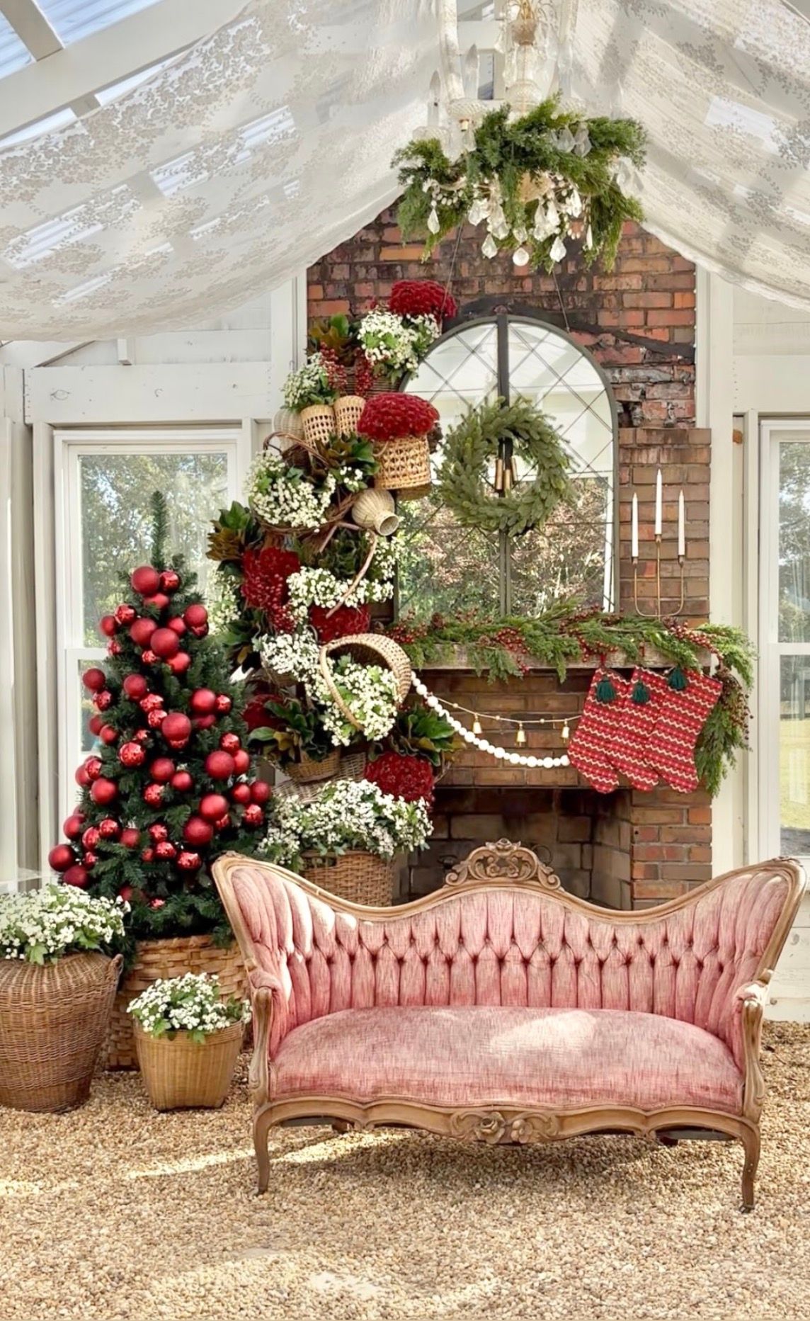 Vintage pink tufted settee in a greenhouse with a red ornament Christmas tree, brick fireplace, wreaths, stockings and holiday greenery.