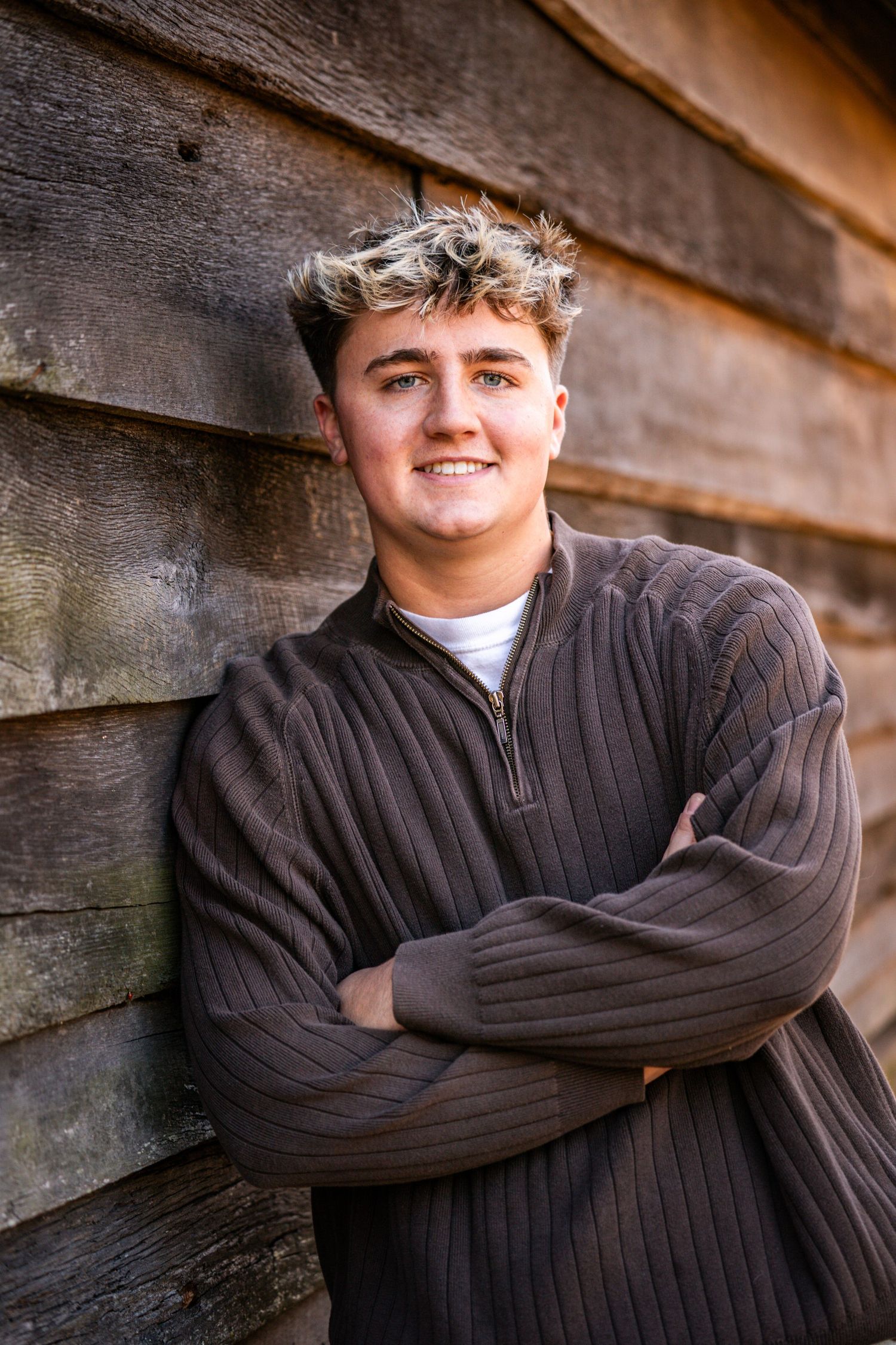 A person wearing a dark ribbed sweater poses against a rustic wooden wall in a casual portrait setting.