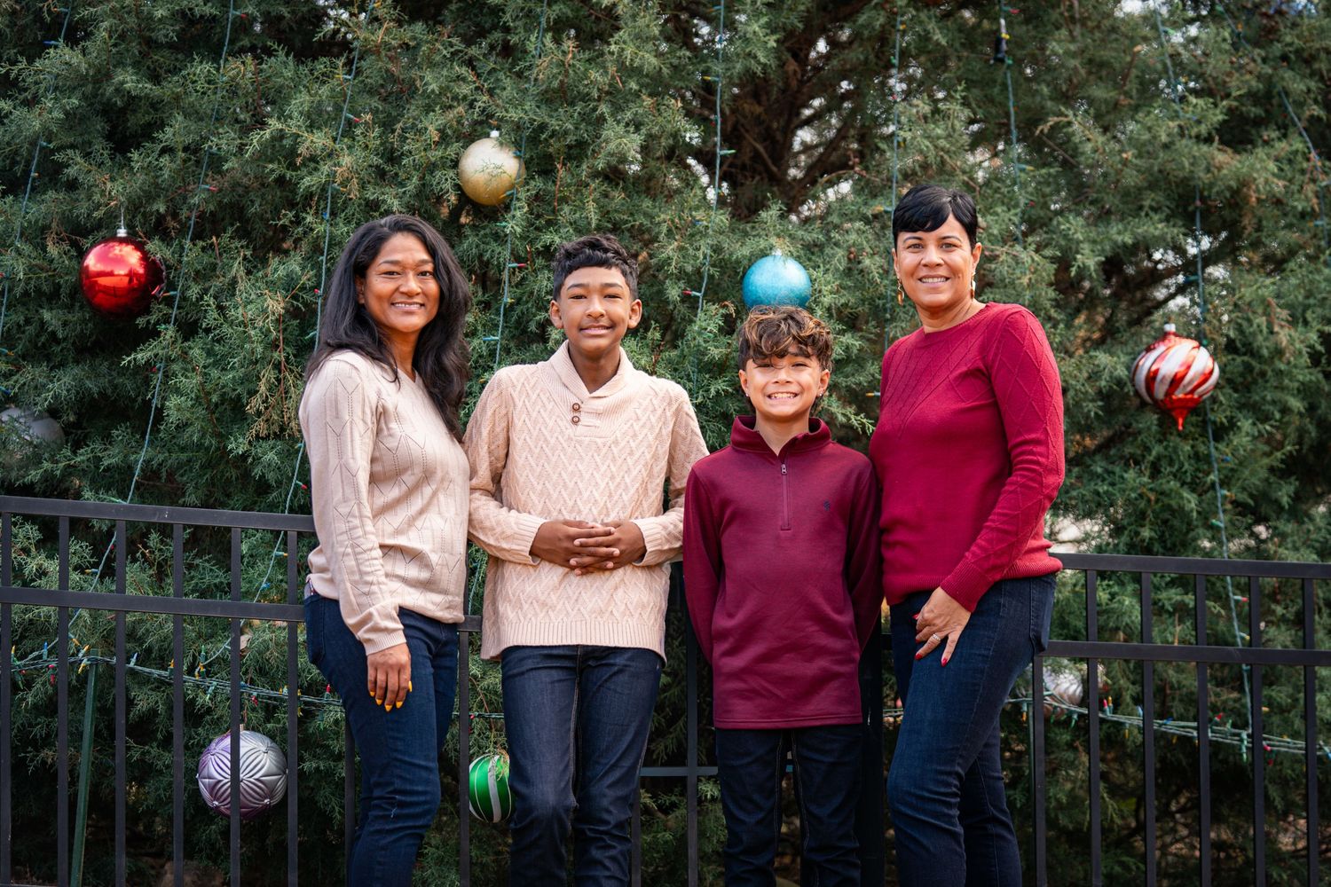 A group of people in casual winter attire stand together smiling in front of an evergreen tree decorated with ornaments.