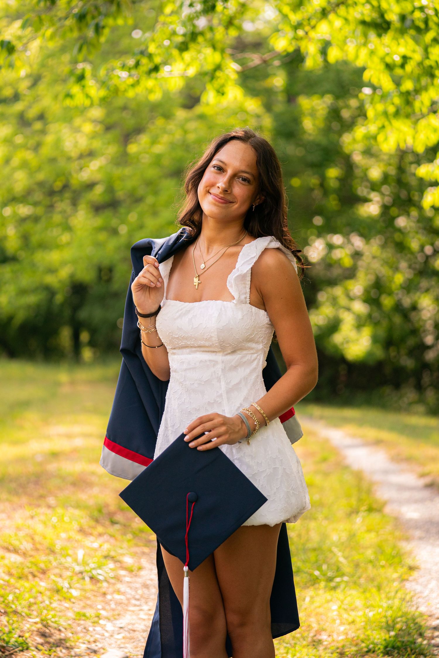 Young woman in white dress holding graduation cap and gown, smiling outdoors on a sunlit tree-lined path.