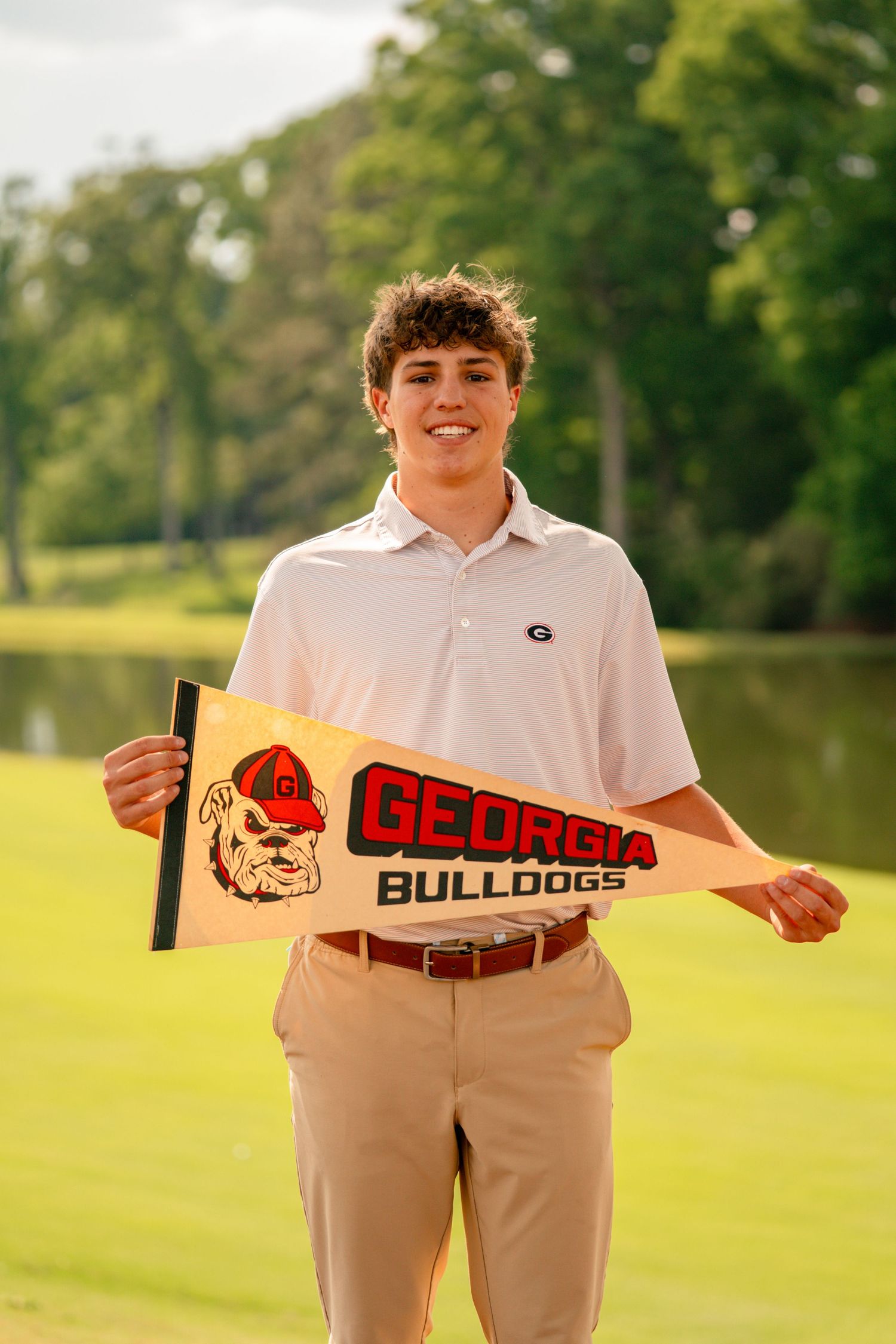 Young man holding a Georgia Bulldogs pennant flag on a golf course with trees and water in background.