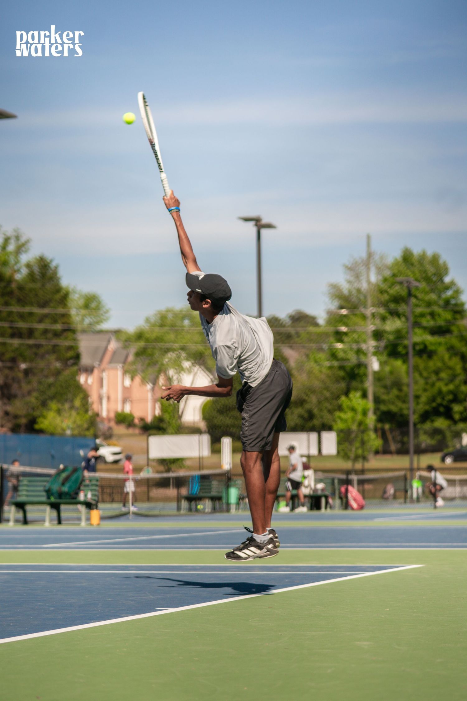 A tennis player executes a powerful serve on an outdoor hard court under a clear blue sky.