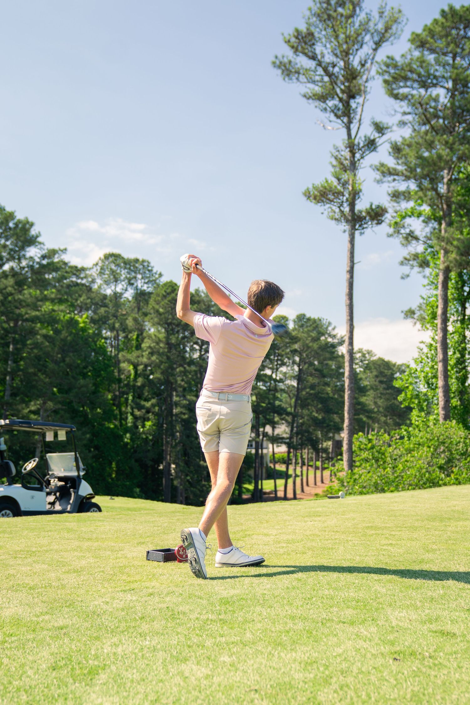 Young male golfer completing a swing on a sunny golf course with trees and a golf cart in the background.