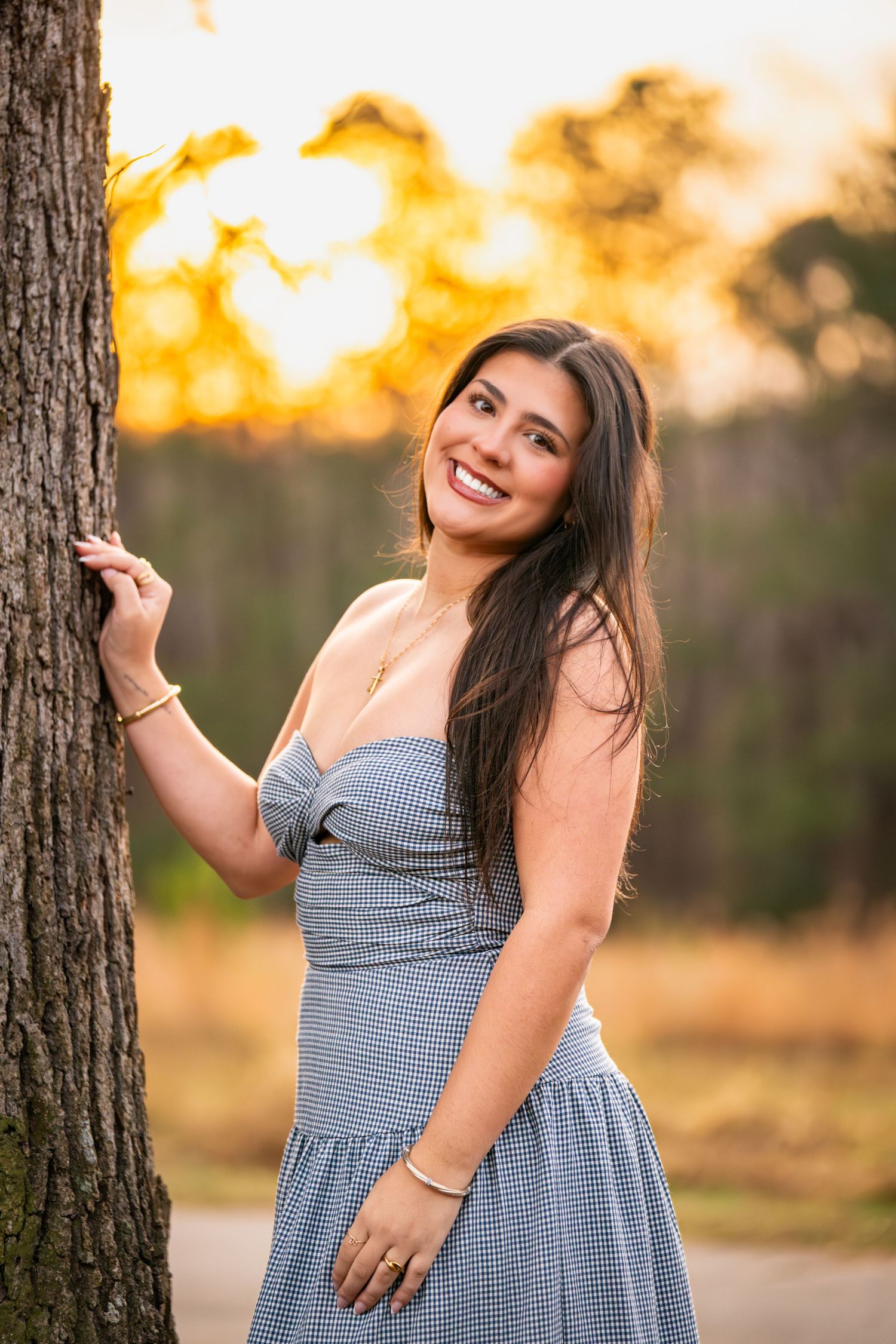 A person in a blue gingham dress poses playfully against a tree trunk during golden hour sunset.