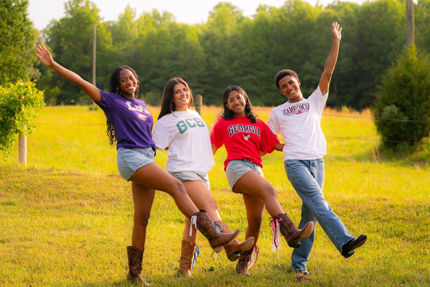 Four college students in university shirts pose joyfully in a green field, arms raised in celebration.