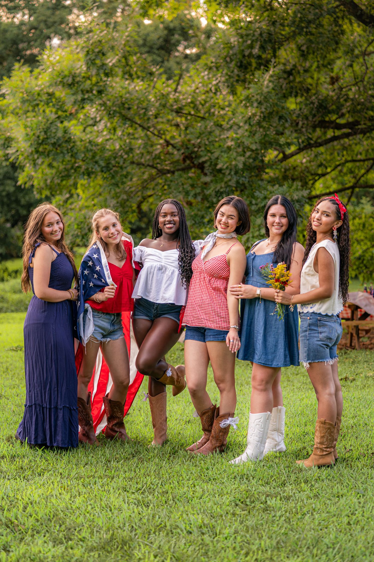 Six diverse young women pose outdoors on green grass, some draped in an American flag, wearing summer outfits and boots.