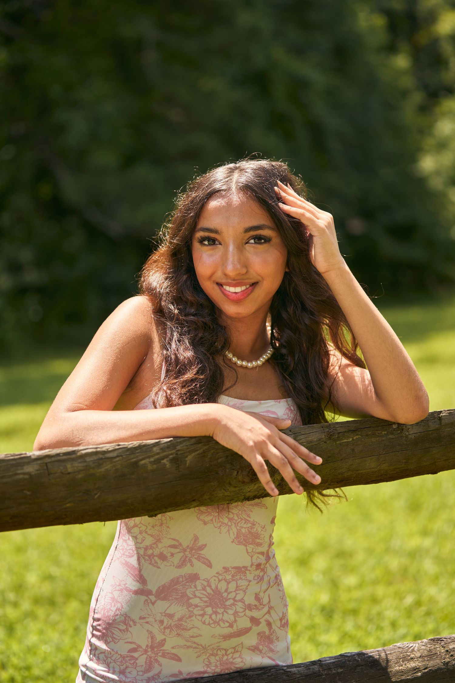 Young woman in floral dress leaning on wooden fence, smiling outdoors with lush green trees in background.