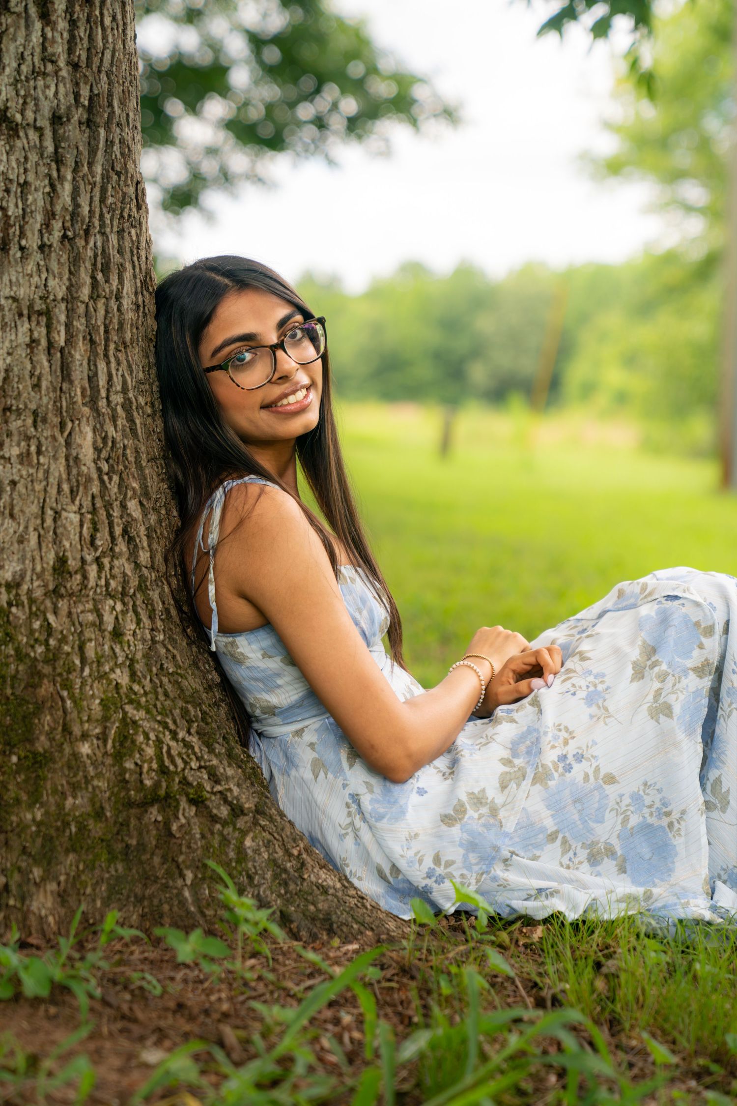 Young woman in floral dress and glasses sitting against a tree in a lush green park, smiling warmly.