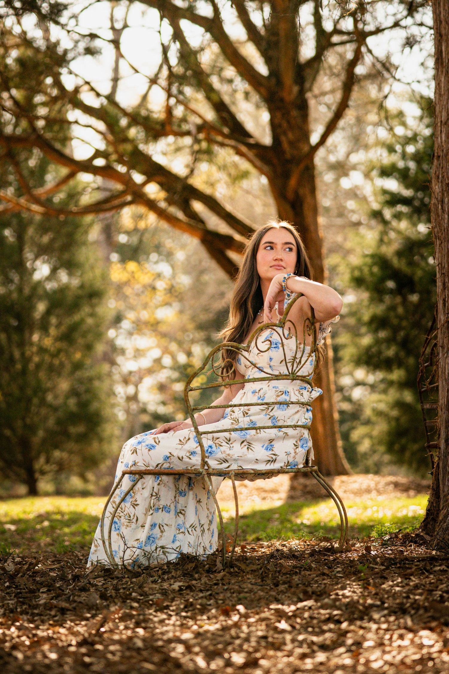 A woman in a floral dress sits on a chair in a wooded area, with sunlight filtering through the trees.