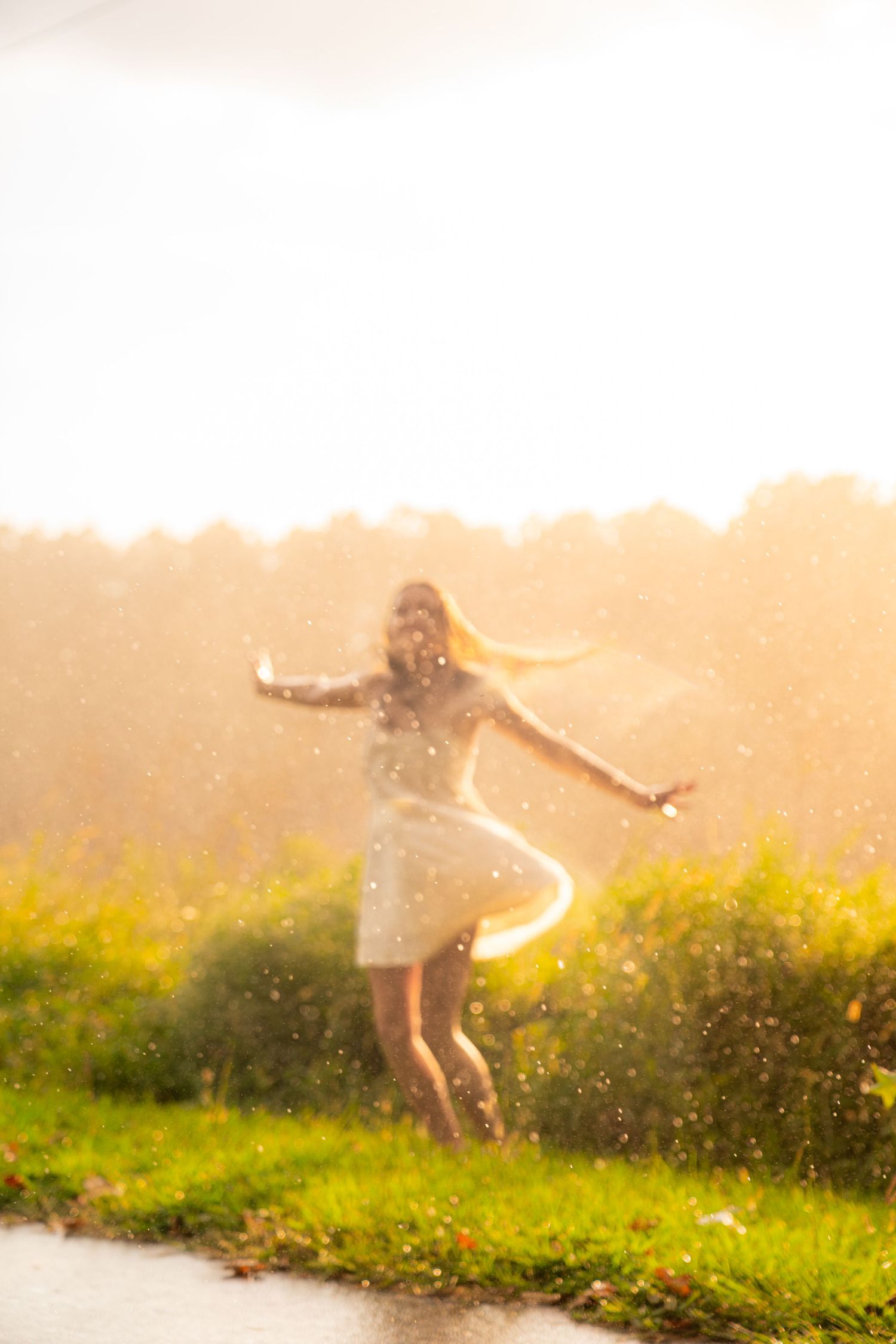 Woman in white dress spinning joyfully in golden sunlight with rain droplets sparkling around her in a lush green field.