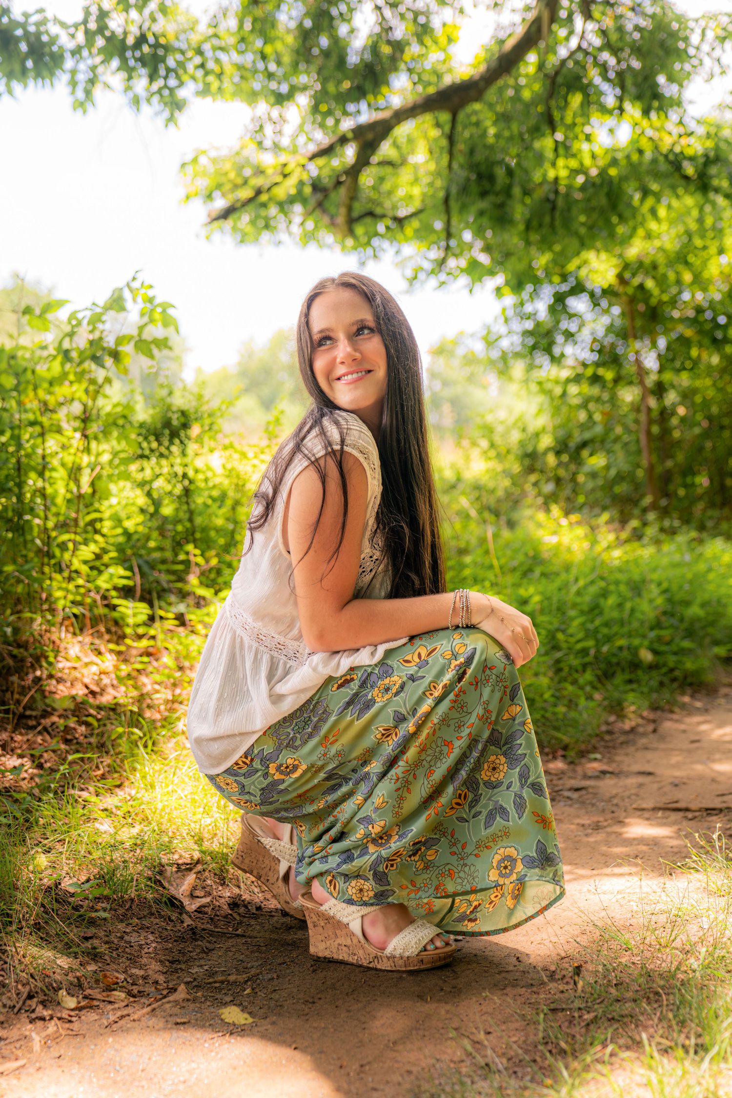 Young woman in floral skirt and white top crouching on a sunlit forest path surrounded by lush green trees.
