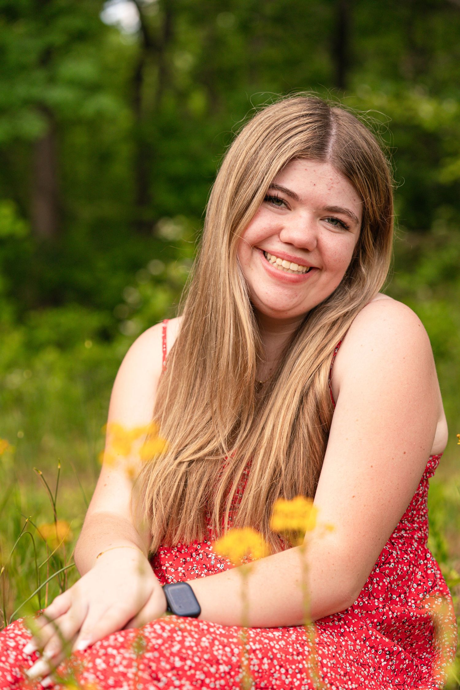 Smiling young woman with long blonde hair wearing a red floral dress, sitting outdoors among yellow wildflowers.