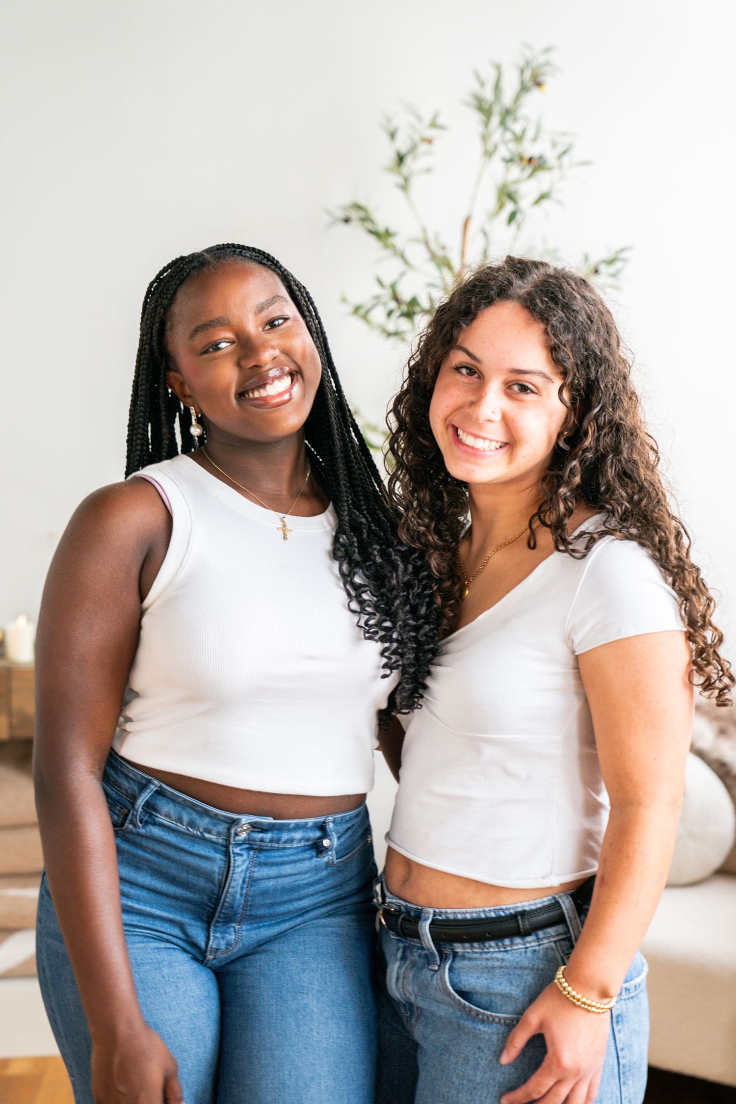 Two young women in white crop tops and jeans smiling together indoors.