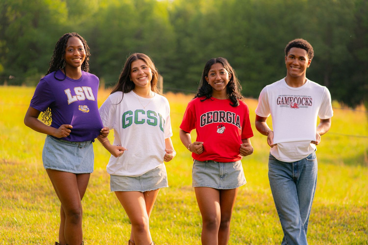 Four college students walk outdoors wearing LSU, GCSI, Georgia, and Gamecocks university shirts in a sunny field.