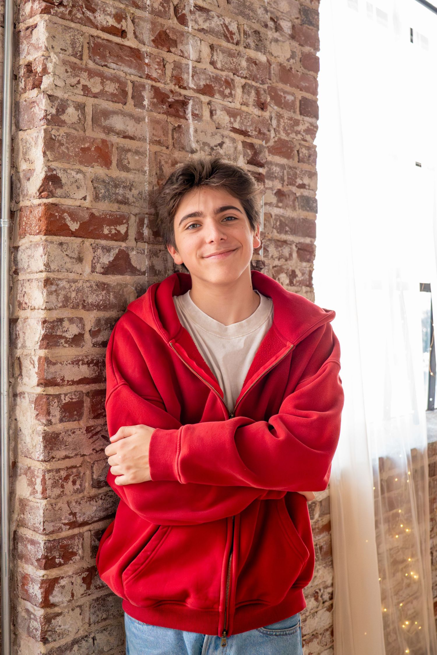 Teen boy in red zip-up hoodie leaning against brick wall, smiling with arms crossed.