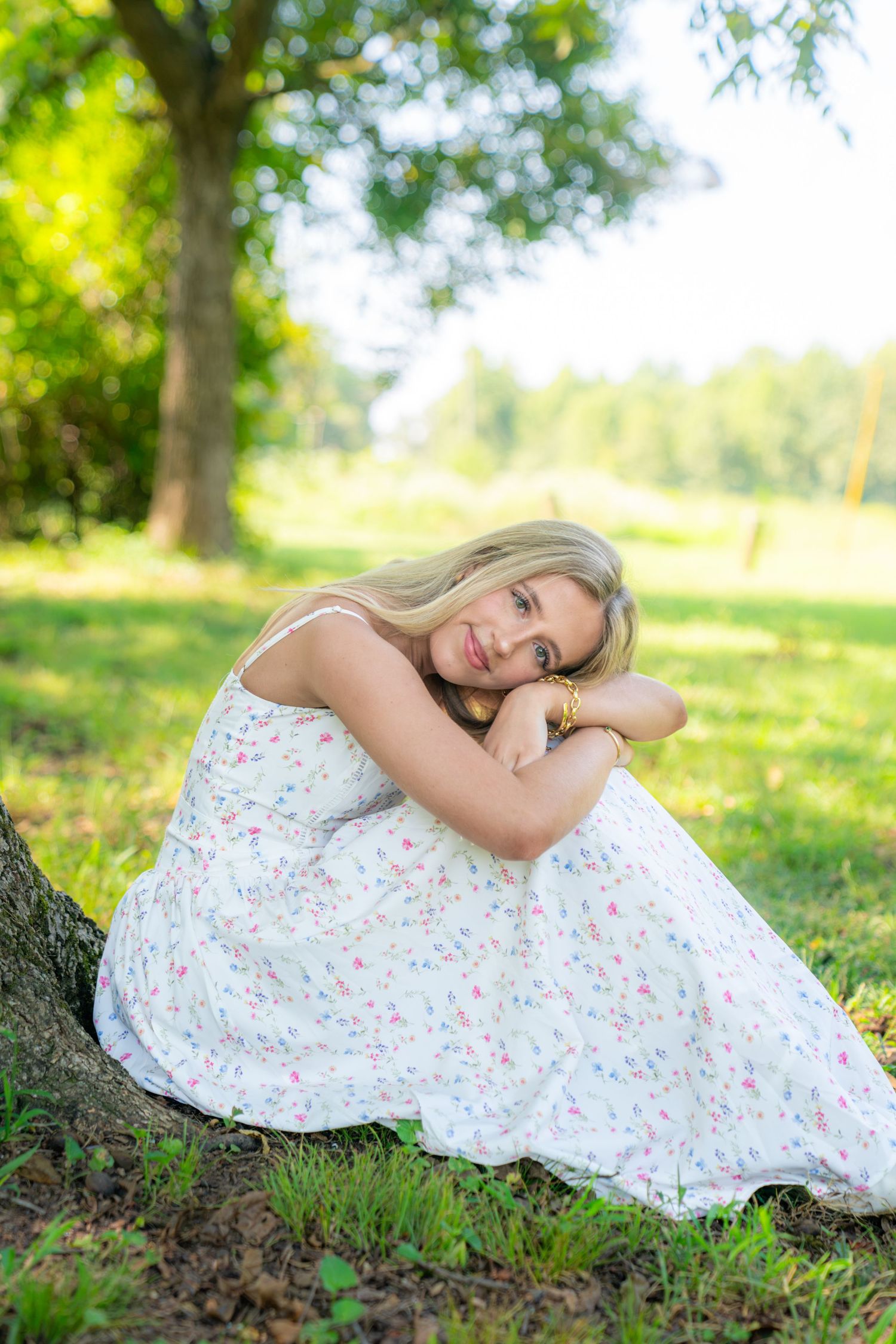 Young woman with long blonde hair in a white floral sundress sitting on green grass beneath a tree, resting her head on her arms.
