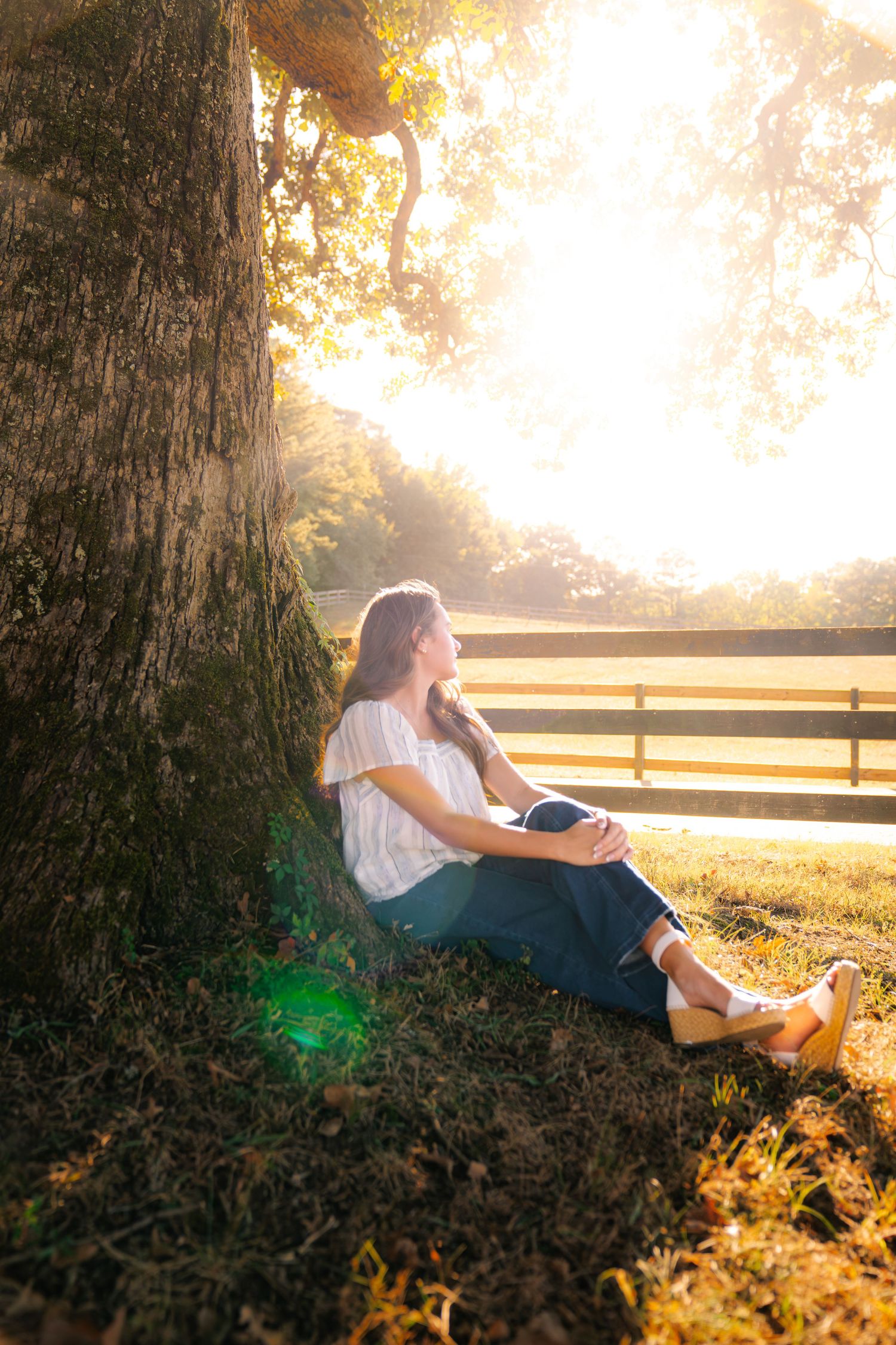 Woman in a white blouse and jeans sits against a large tree trunk, gazing at the glowing sunset beside a wooden farm fence.