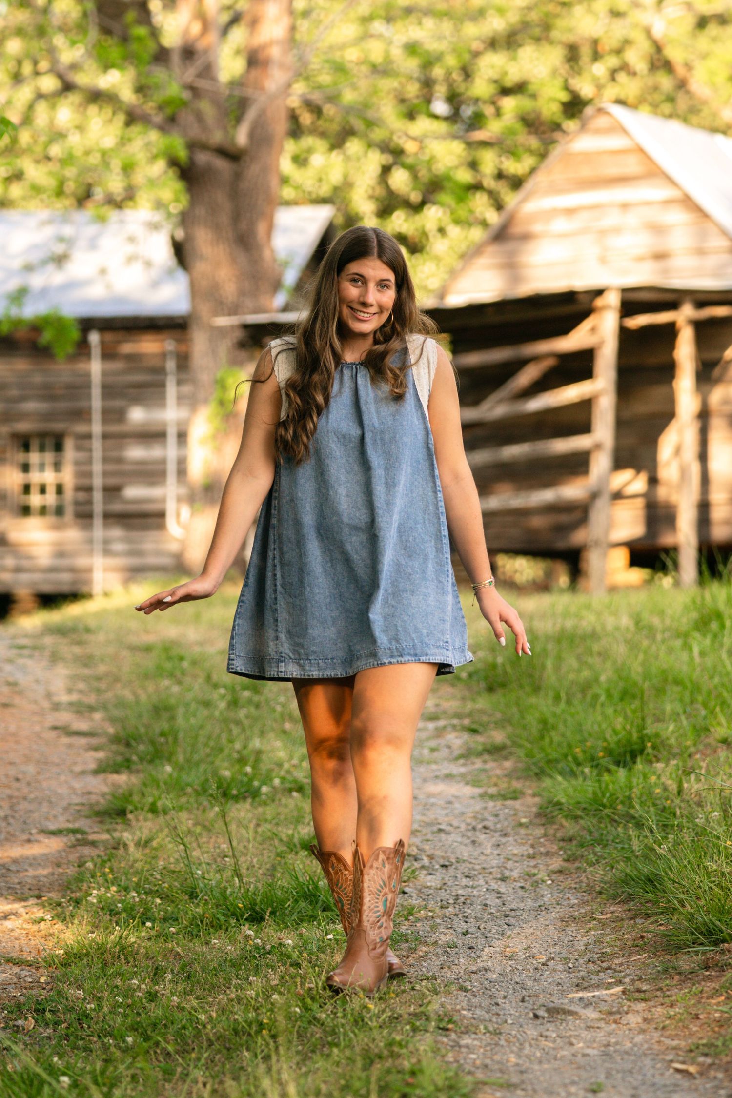 Young woman in denim dress and cowboy boots walking on a path near a rustic wooden barn in a green field.