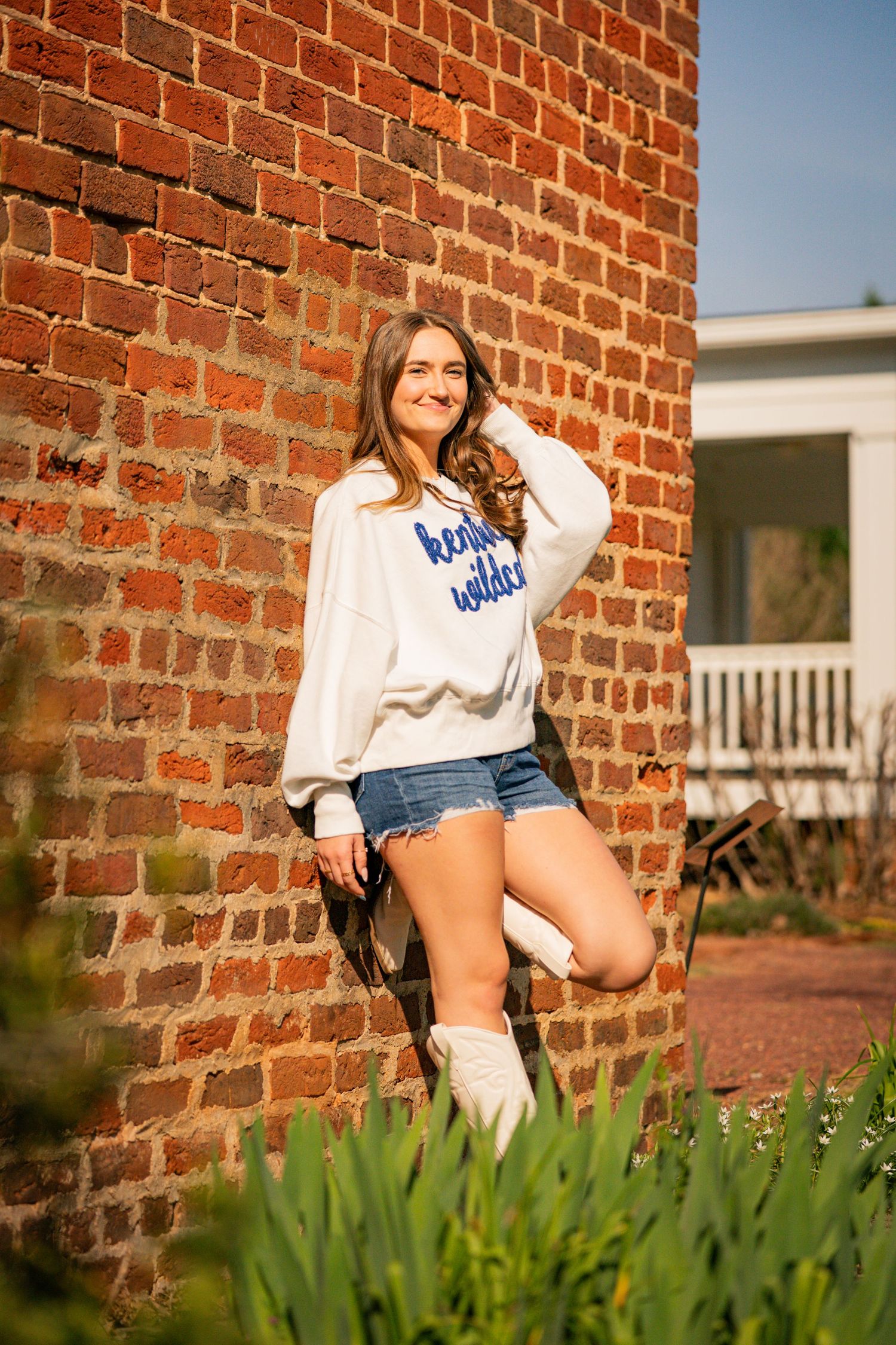 A young woman with long brown hair is leaning against a brick wall, wearing a white sweater and denim shorts.