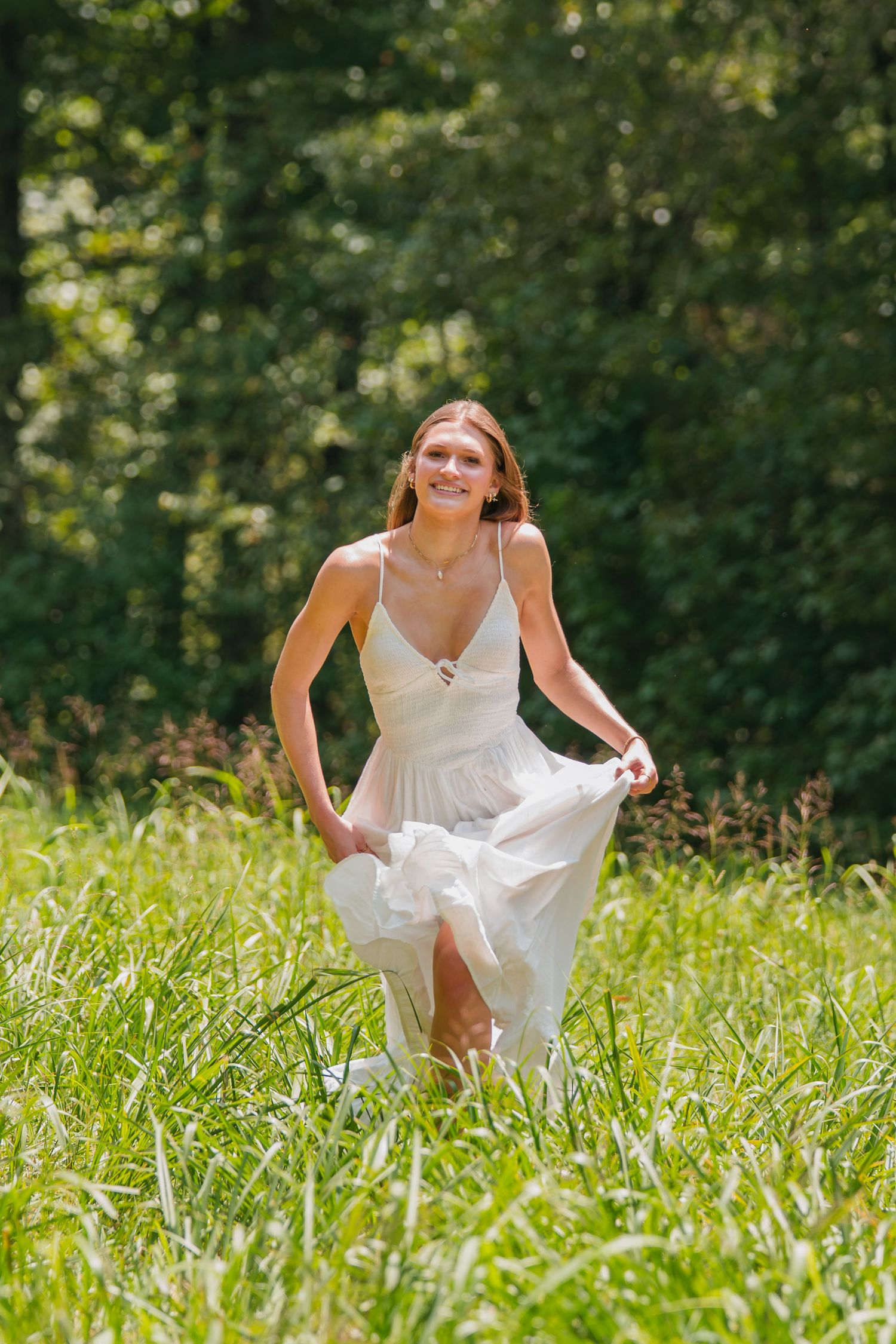 Young woman in white dress walking through green meadow with forest background on sunny day.