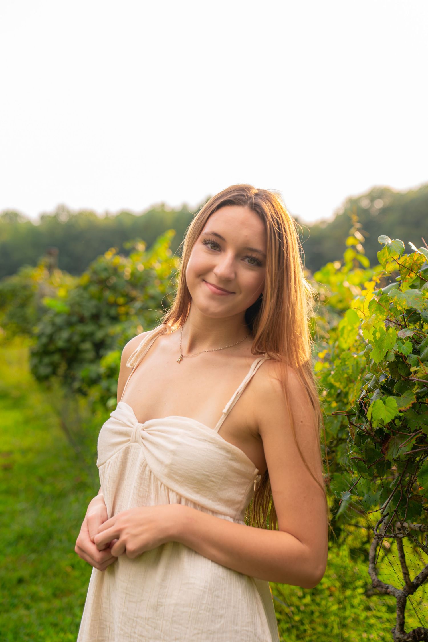Young woman in white sundress smiling in lush green vineyard during golden hour.