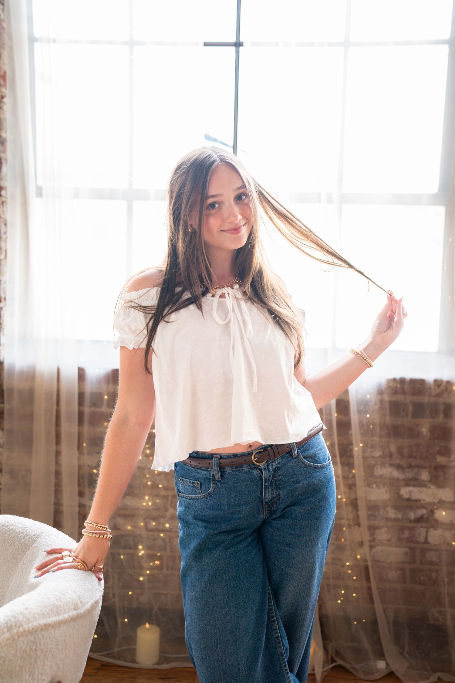 Young woman in white crop top and jeans smiling near bright window, playing with her hair in a cozy indoor setting.