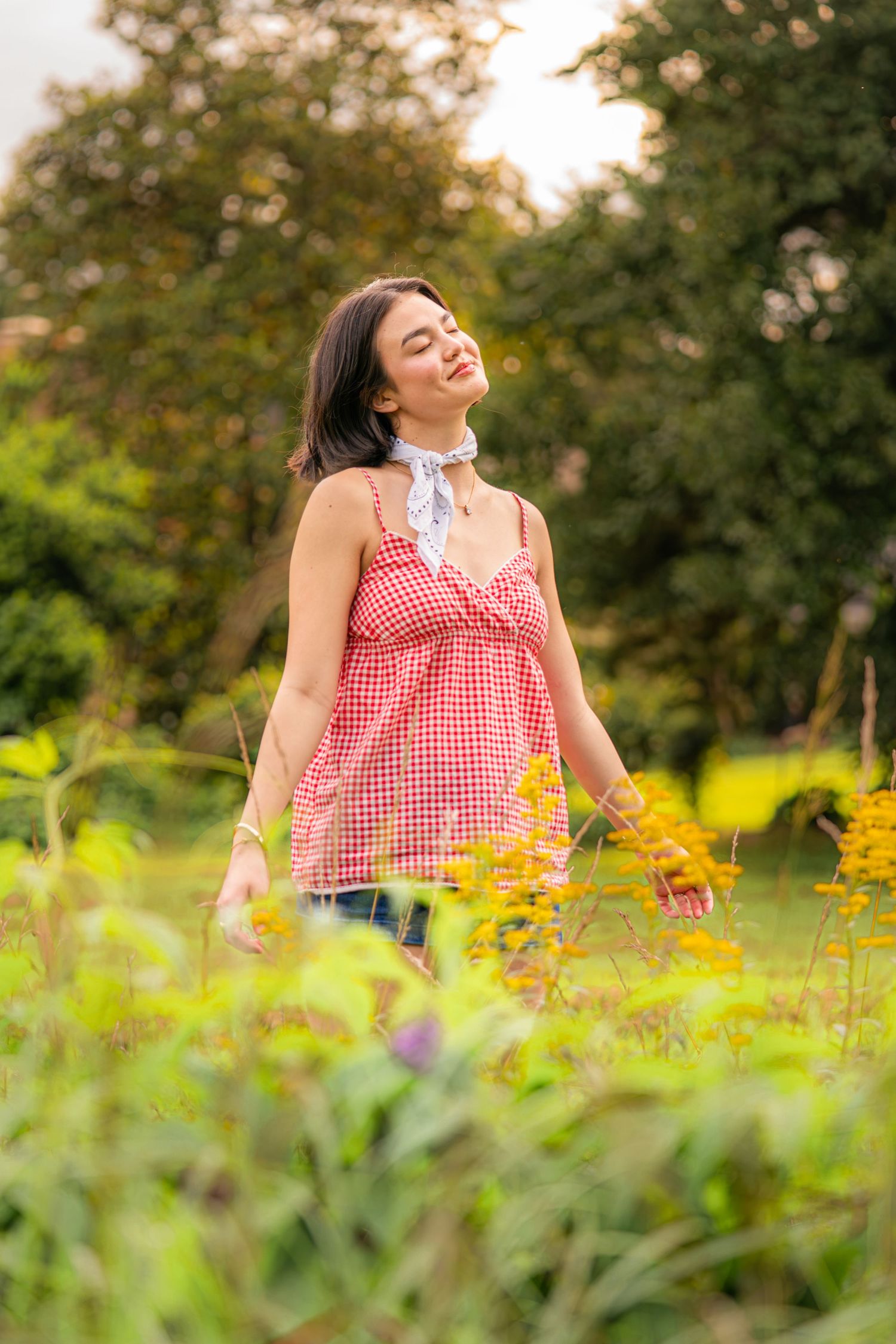 Woman in red gingham top standing in a lush green meadow with wildflowers, eyes closed, enjoying nature.