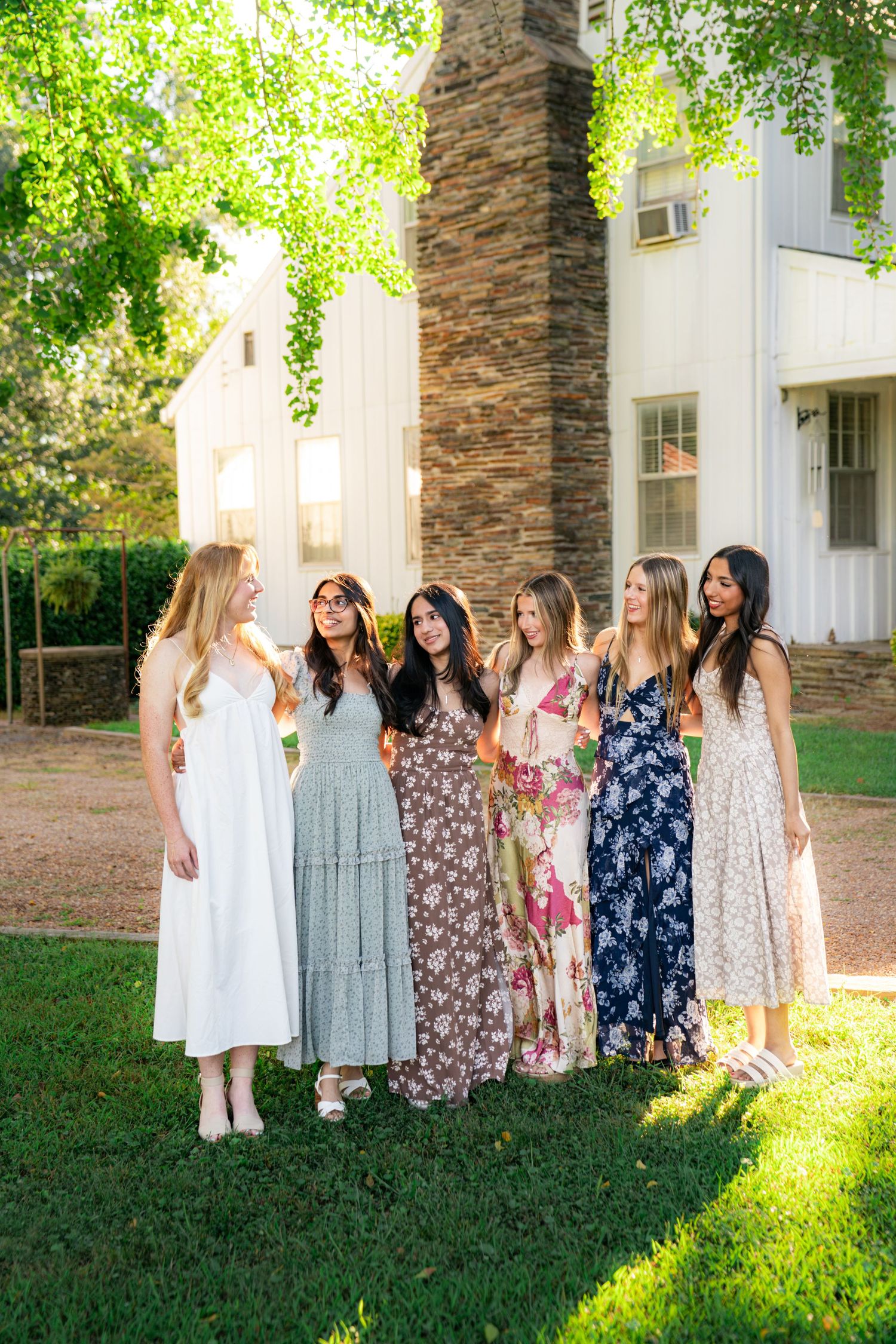 Six women in colorful floral and pastel sundresses stand arm in arm, smiling at each other on a sunlit lawn in front of a white house.