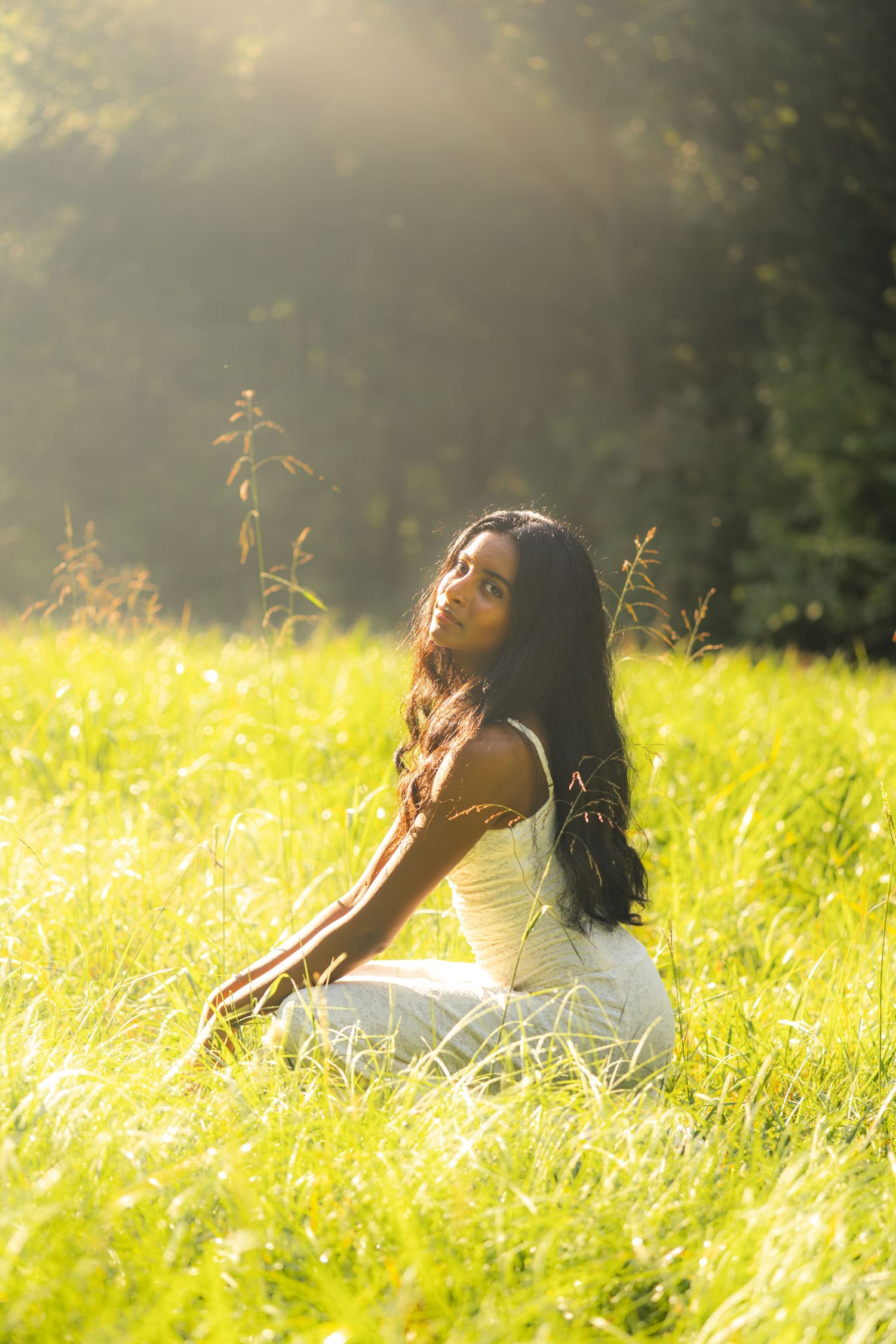 Young woman in white dress sitting in sunlit green meadow with long dark hair, surrounded by tall grass and trees.