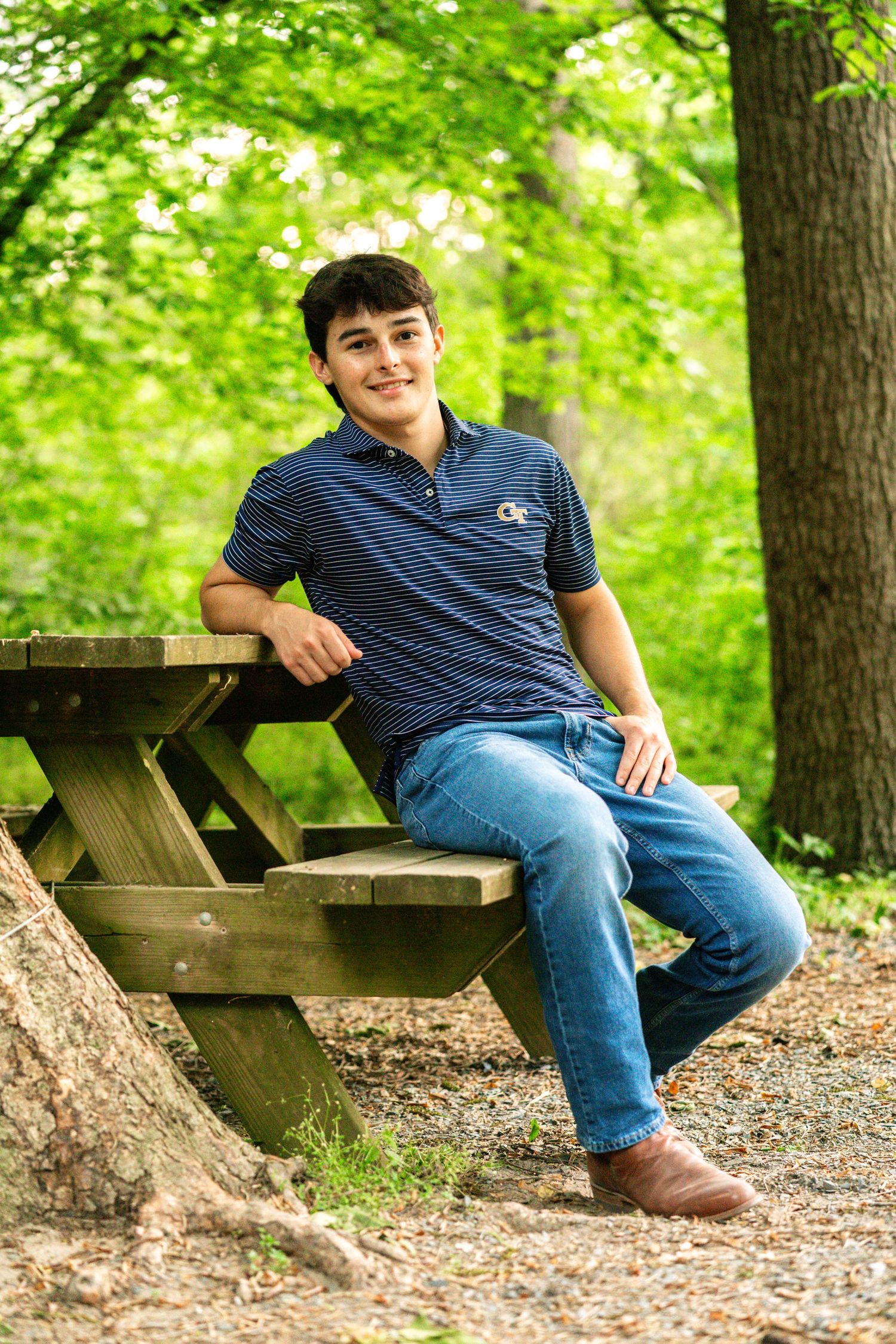 Young man smiling in navy striped polo sits at wooden picnic table in lush green park setting.