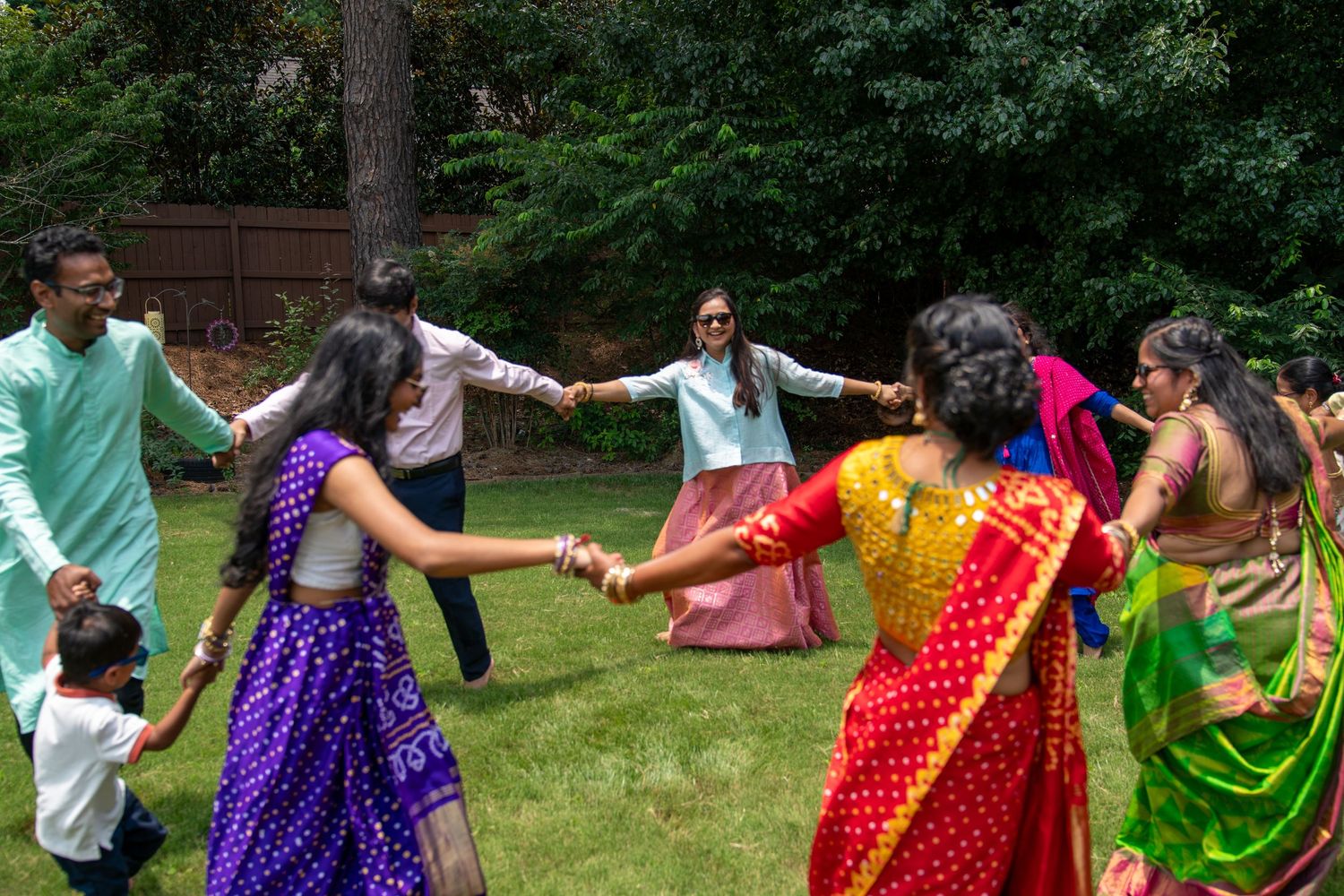 Group of people in colorful traditional Indian attire dancing in a circle outdoors on a sunny day.