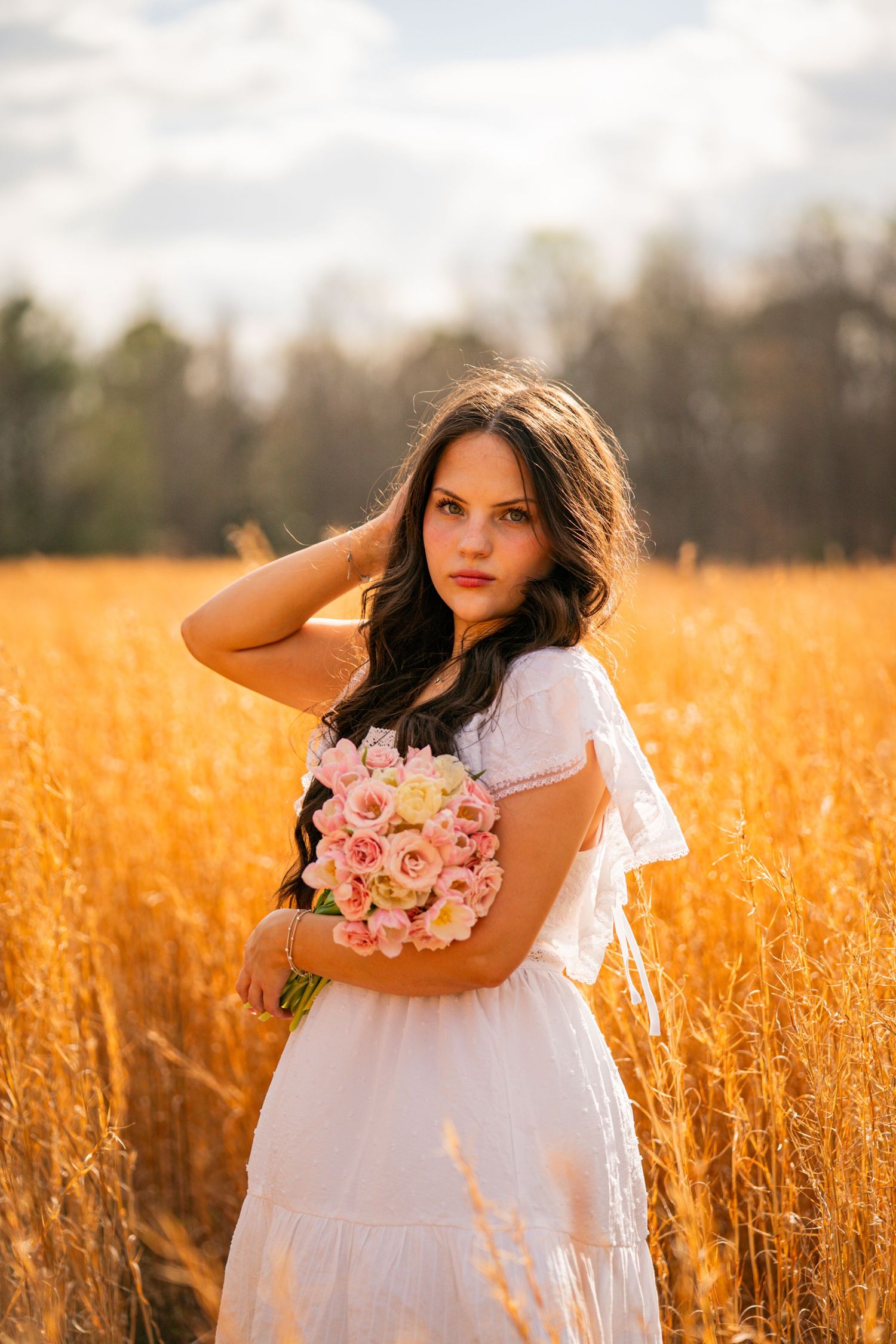 A person in a white dress holds pink roses while standing in a golden wheat field at sunset.