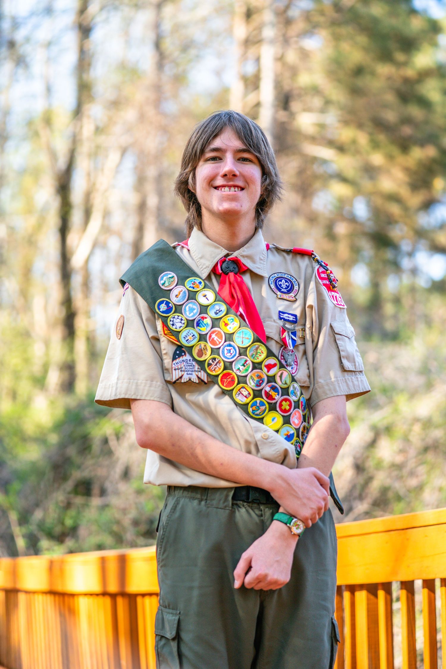 A young person in a Scout uniform with many badges and patches on their shirt, standing outdoors near a wooden fence.
