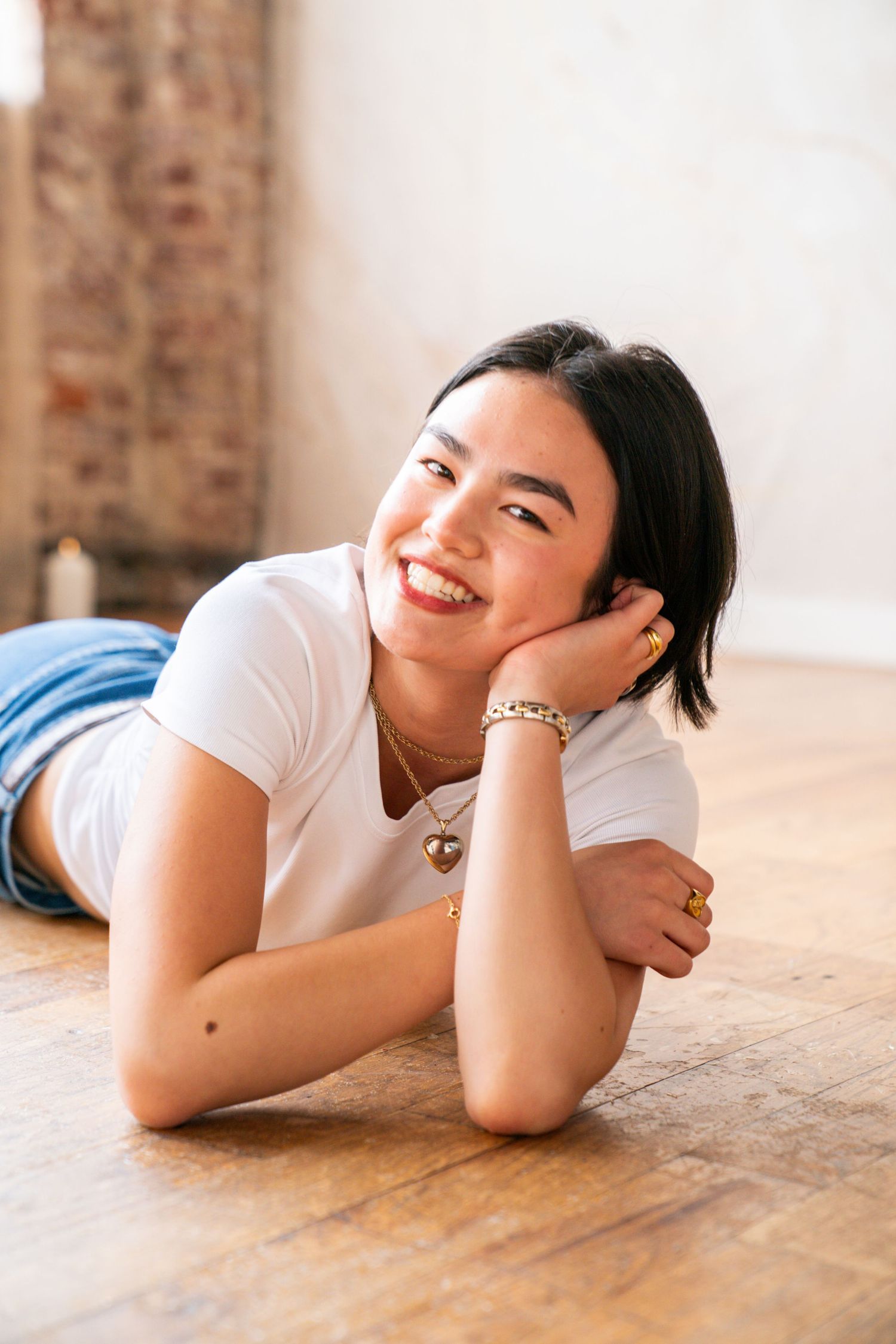 Smiling young woman lying on wooden floor wearing white t-shirt and jeans, adorned with gold jewelry.