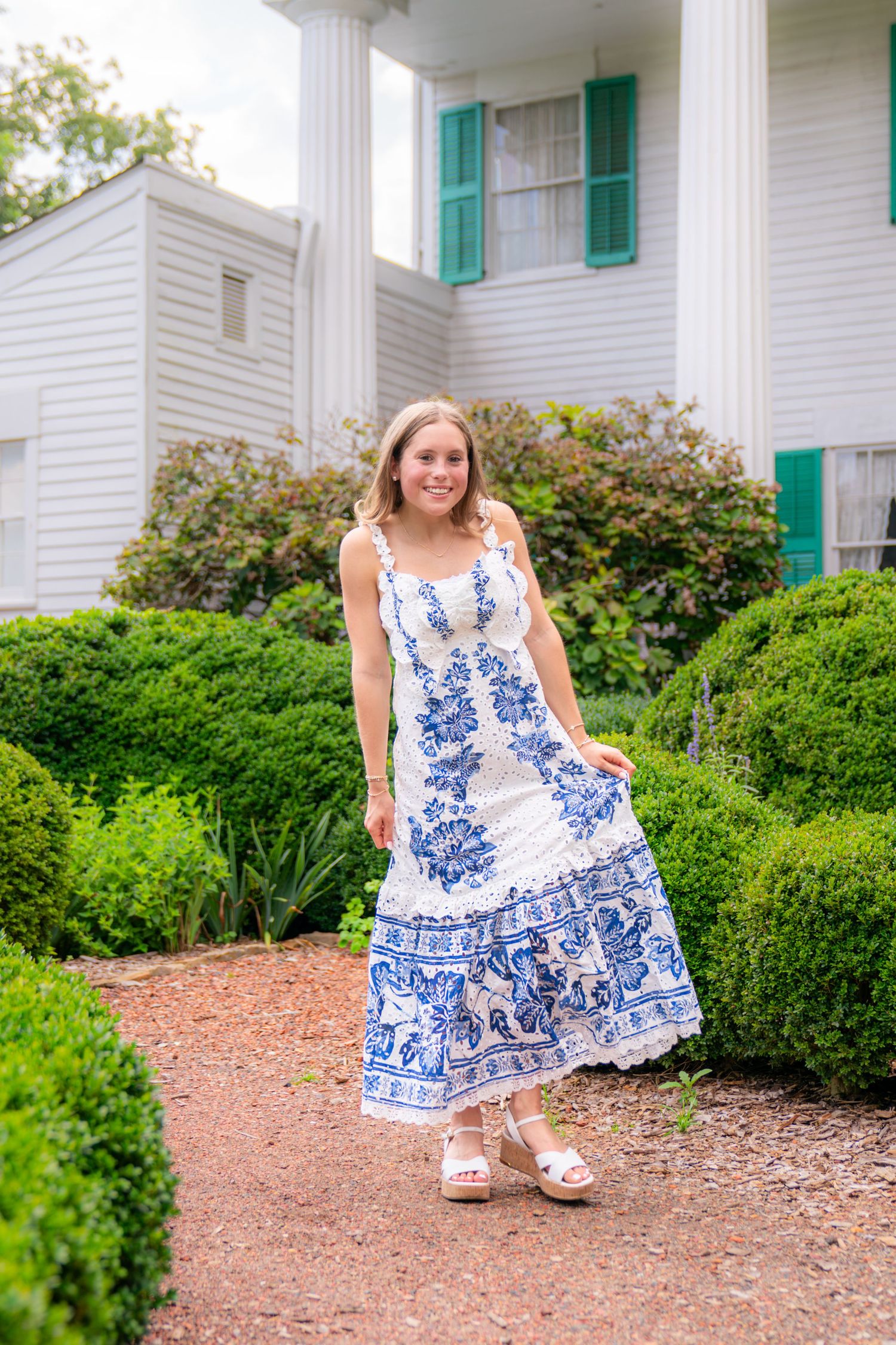 Young woman in white and blue floral maxi dress posing in garden path before white colonial house with green shutters.