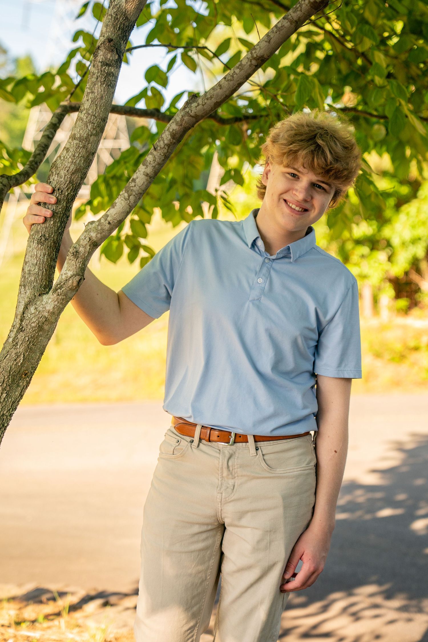 Smiling teenage boy in blue polo shirt and khaki pants leaning against a tree outdoors on a sunny day.
