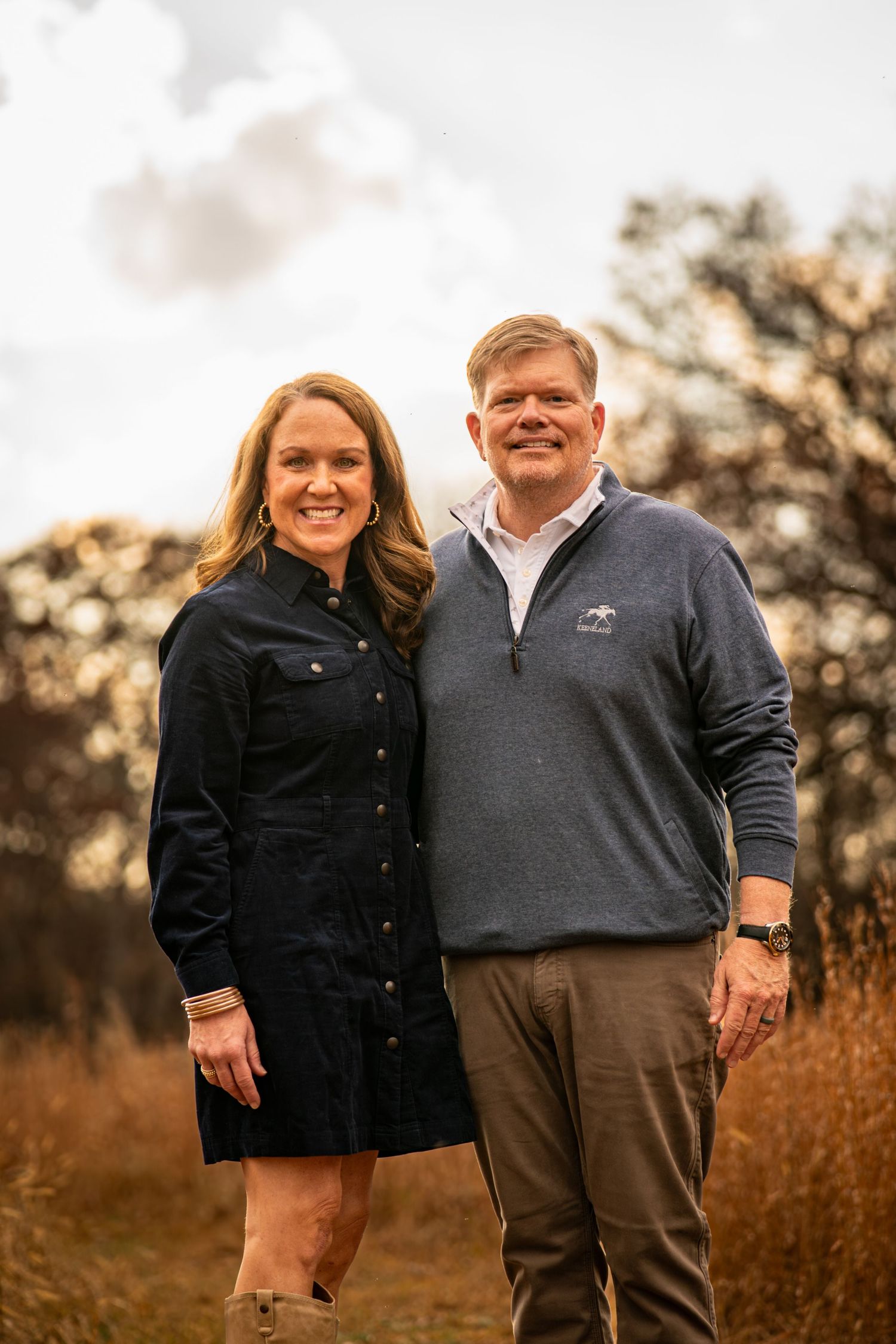 A couple in coordinating black and gray outfits pose together in a golden field at sunset.