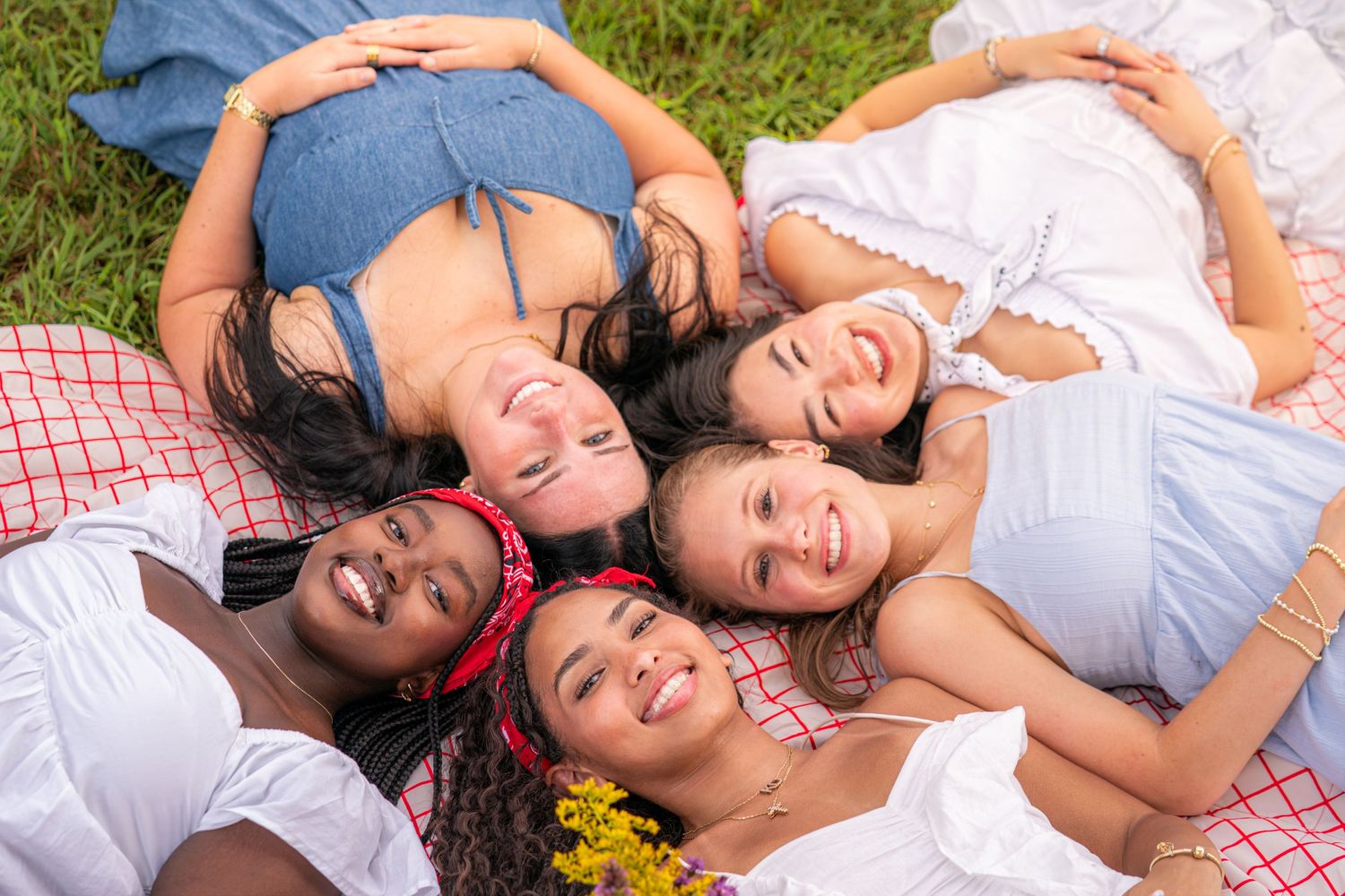 Five diverse young women smiling and lying together on a picnic blanket outdoors, heads together in a circle.