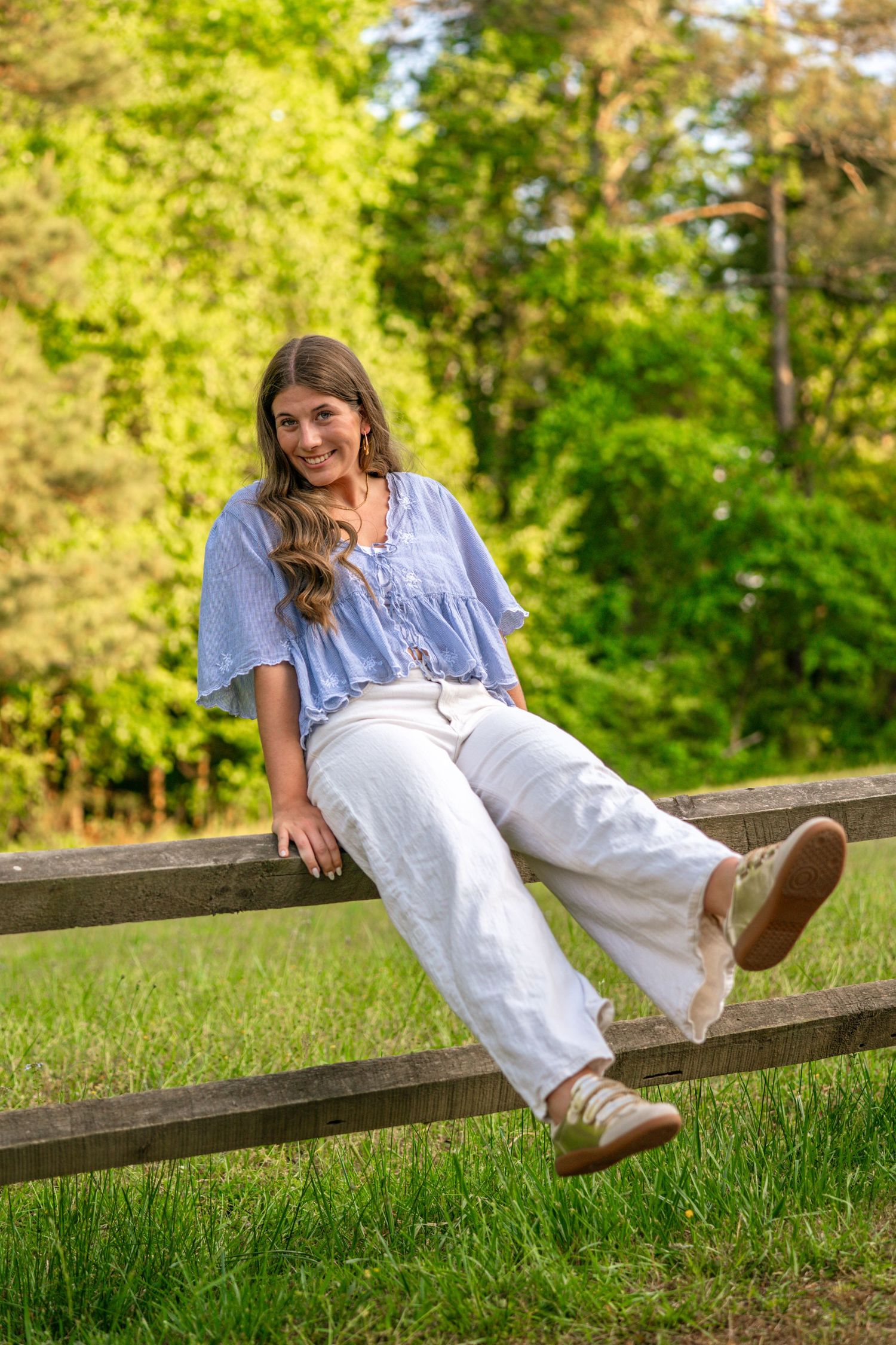 A smiling young woman in a blue top and white pants sits relaxed on a wooden fence surrounded by lush green trees.