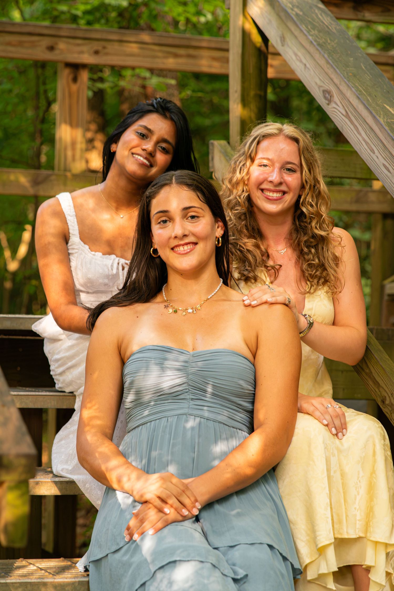 Three smiling young women in dresses pose together on wooden outdoor steps surrounded by lush green trees.