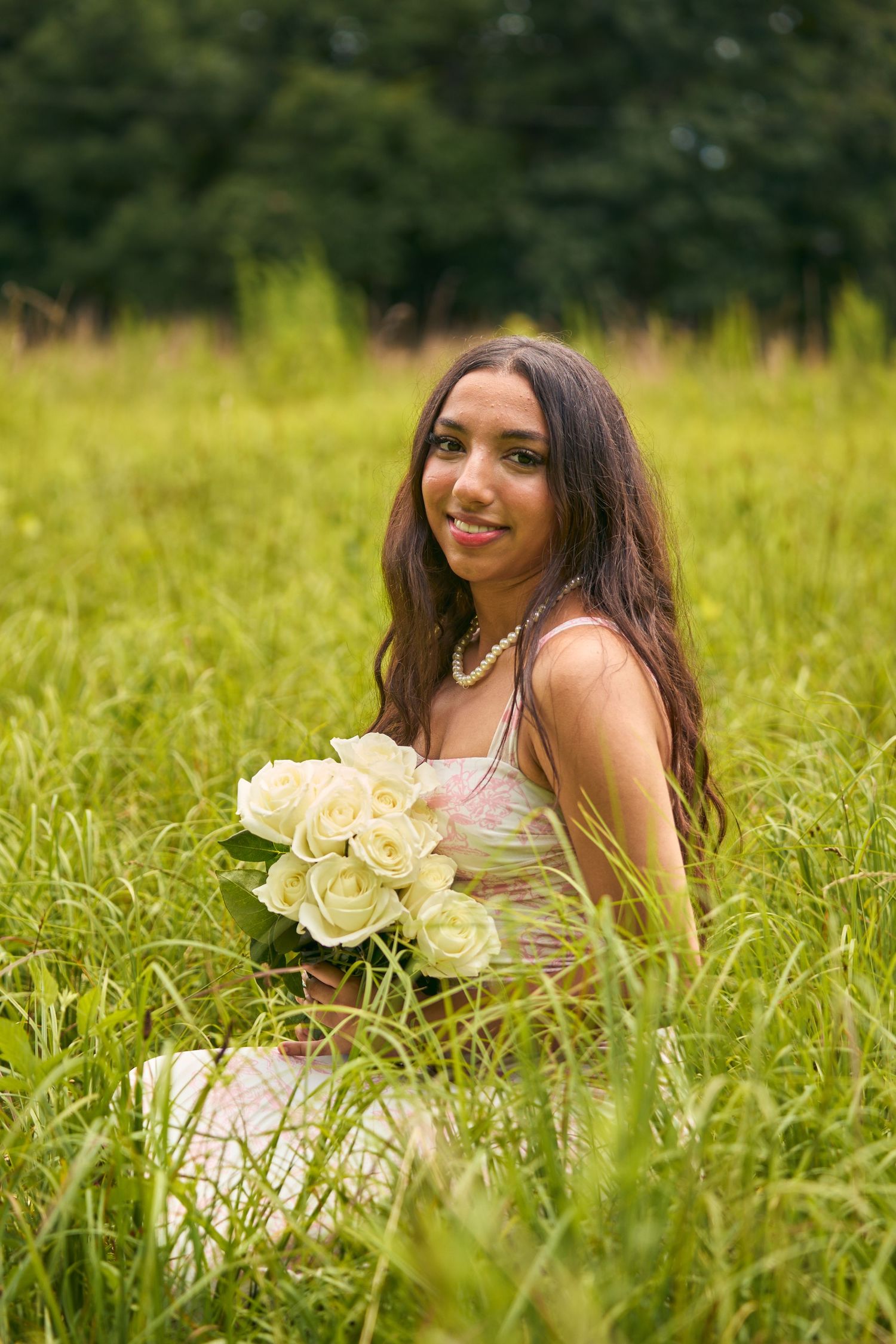 Young woman sitting in tall green grass holding white roses bouquet, wearing floral dress and pearl necklace, smiling outdoors.