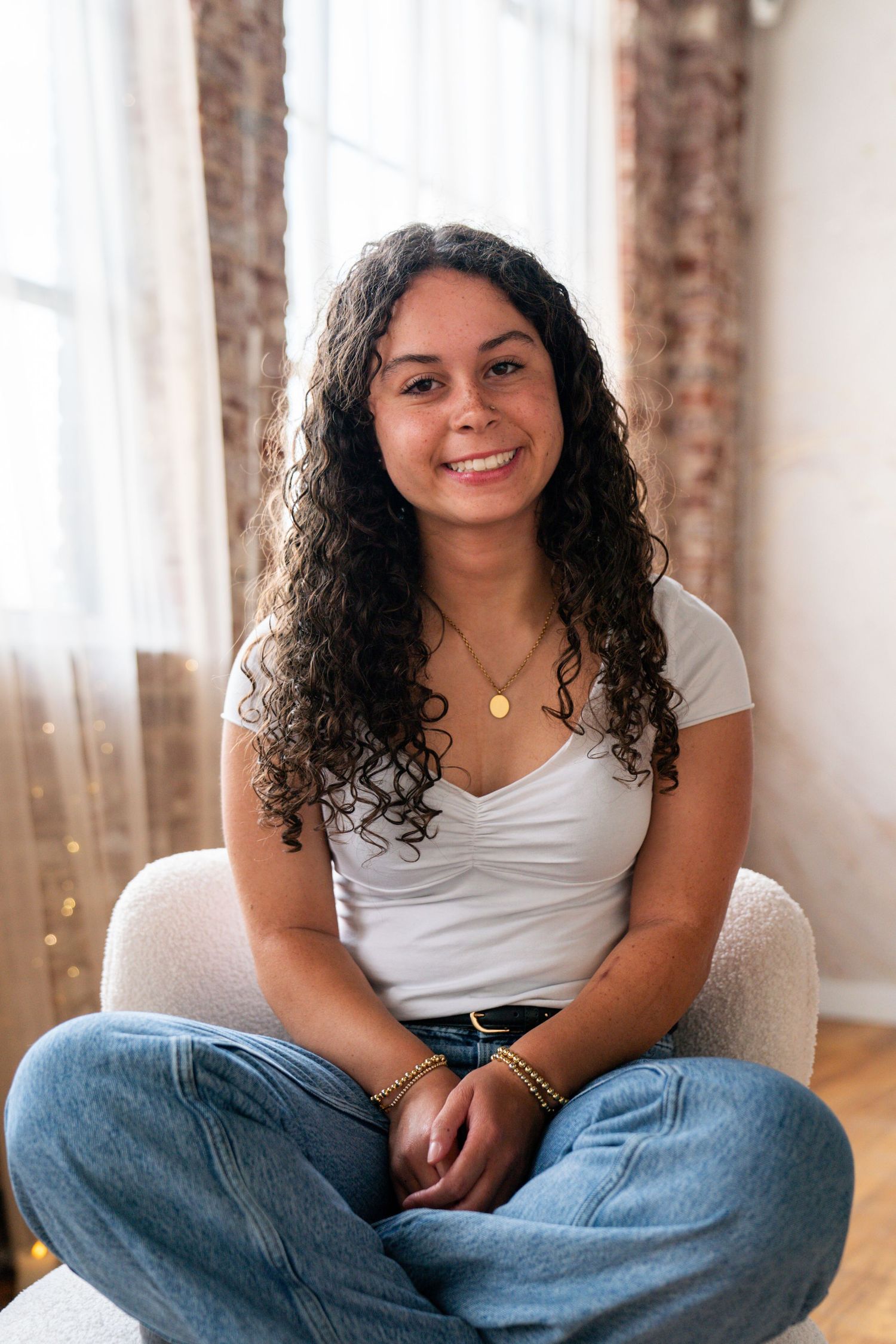 Young woman with curly hair sitting cross-legged in a chair, smiling, wearing a white top and jeans.