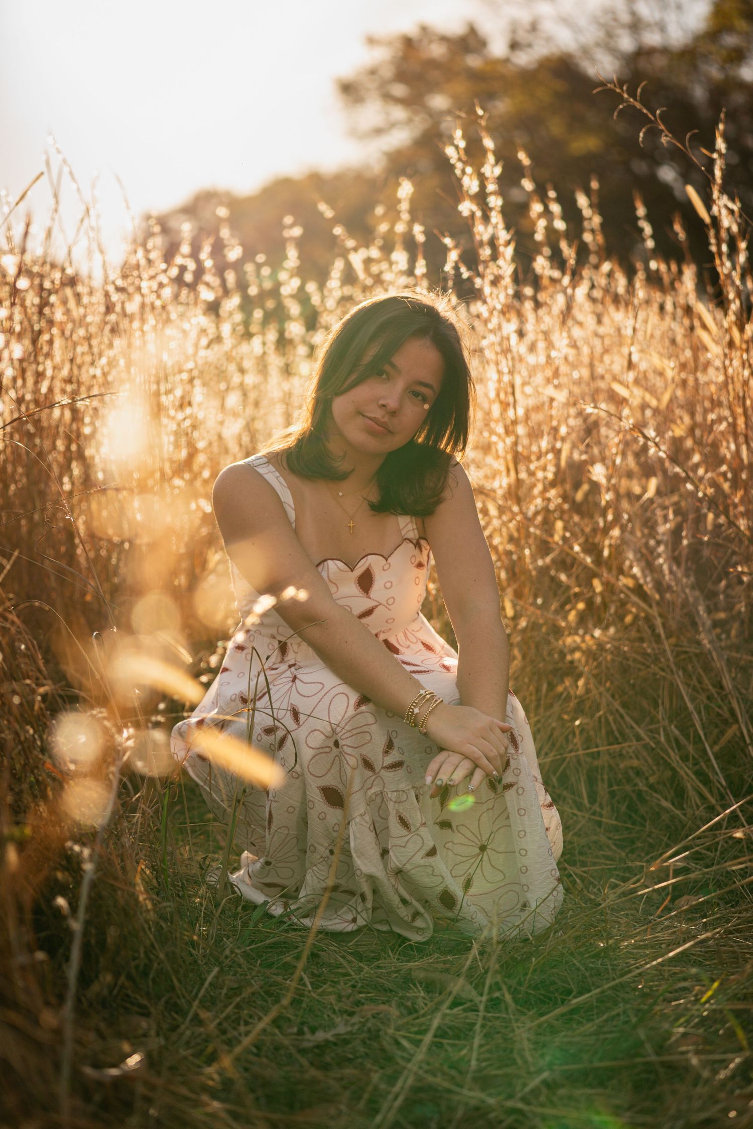 Dreamy golden hour portrait of someone in a white floral dress sitting in a sun-lit meadow of tall grass.