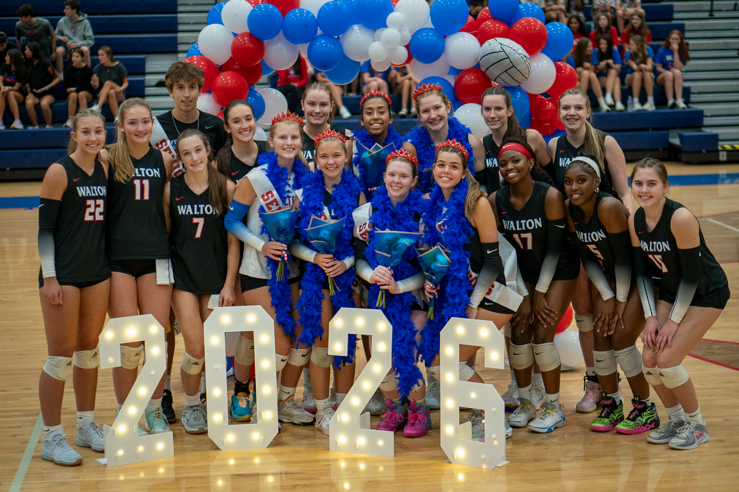 Sports teams gather for group photo under balloon arch in school gymnasium.