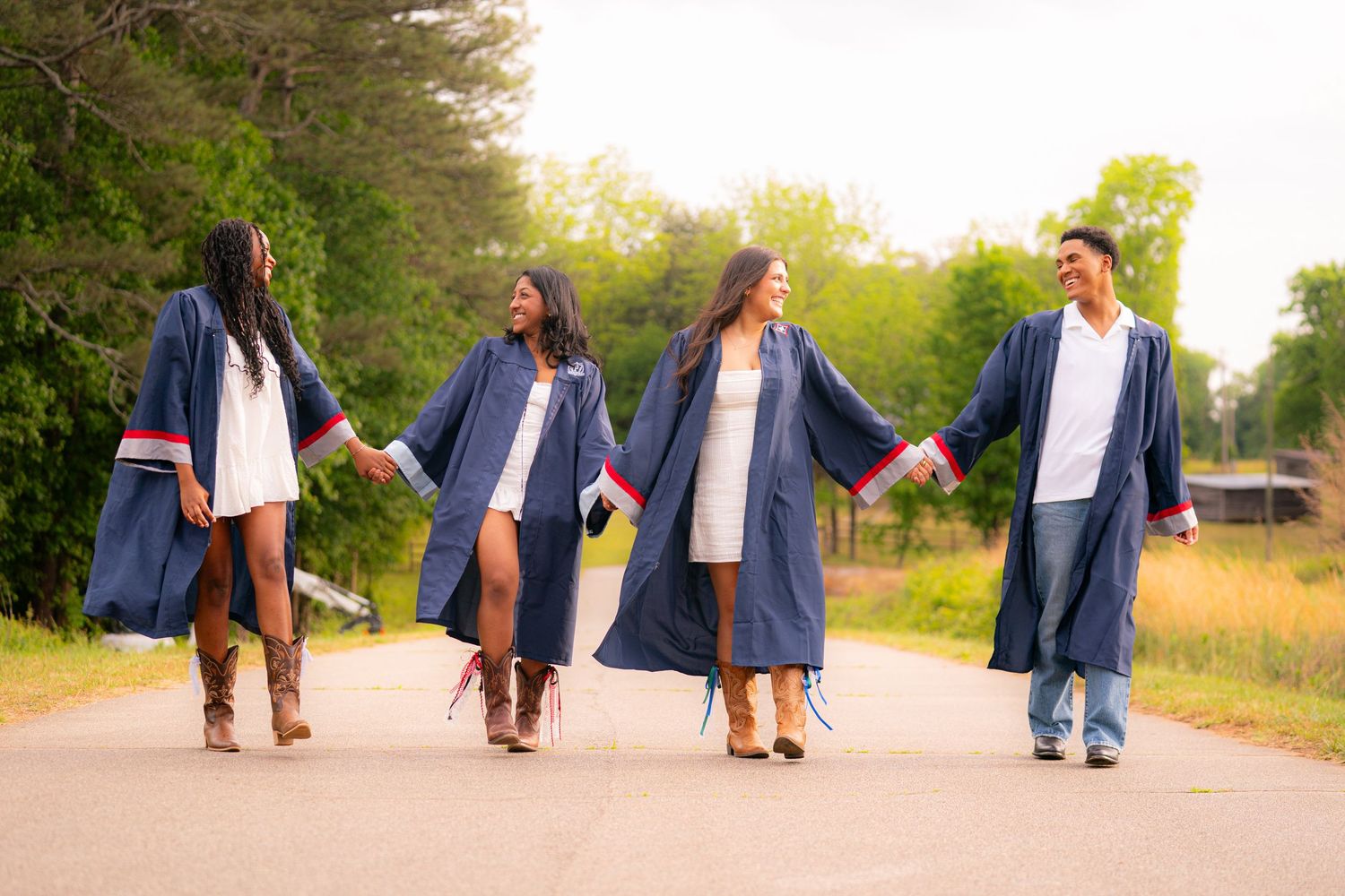 Four graduates in navy blue gowns walk hand-in-hand on a tree-lined road during an outdoor graduation photo session.