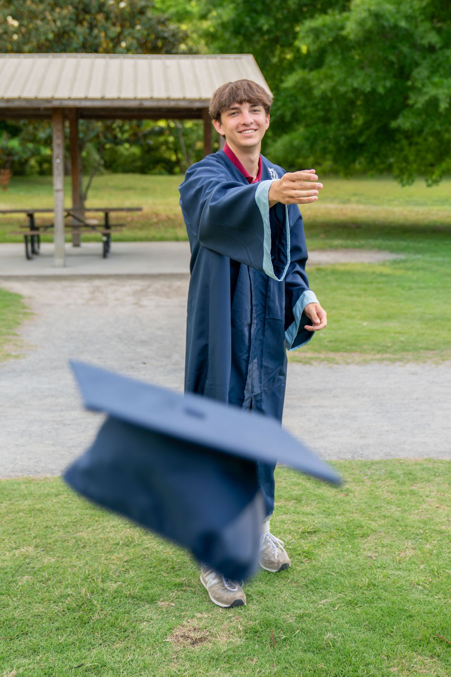 Smiling graduate in blue gown tossing mortarboard cap in the air outdoors in a green park setting.