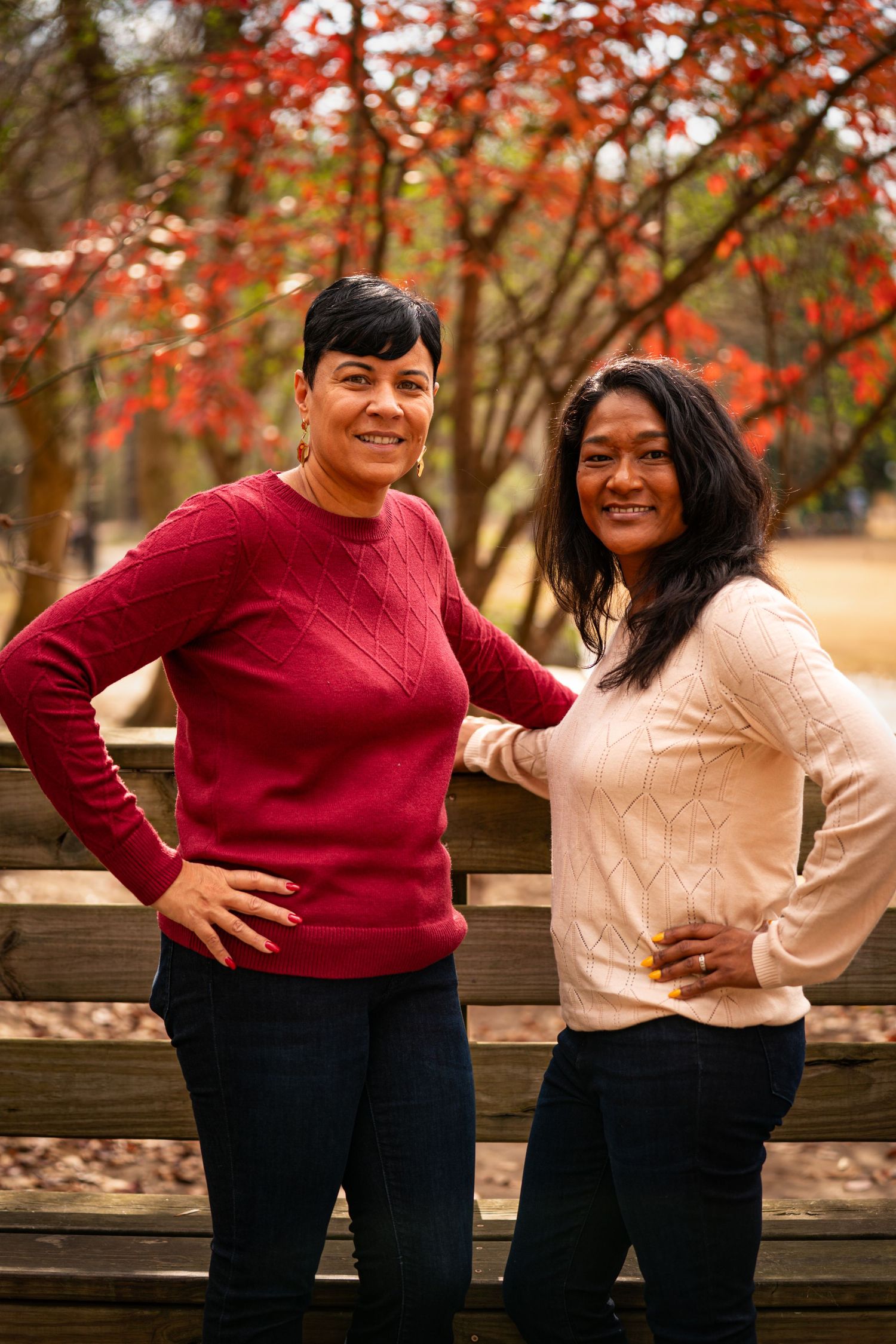 Two women smiling outdoors in fall, wearing red and cream sweaters, posing near a wooden fence with autumn foliage.