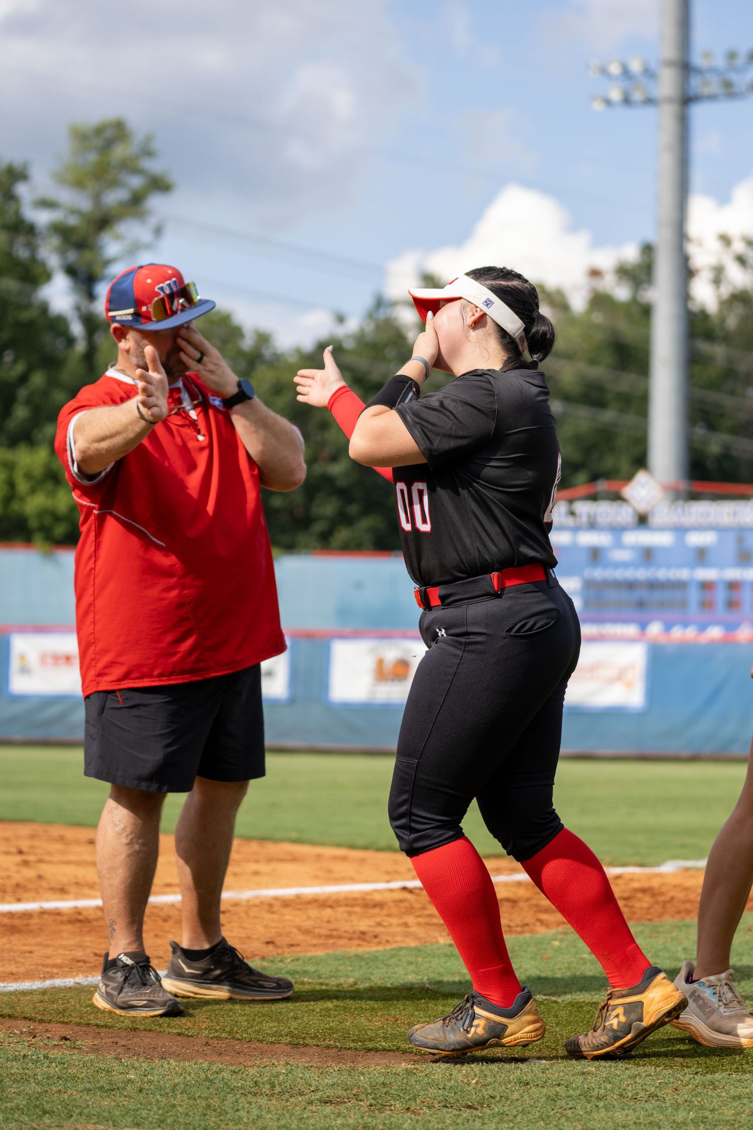 Softball player #00 in black uniform high-fives coach in red at base during outdoor game.