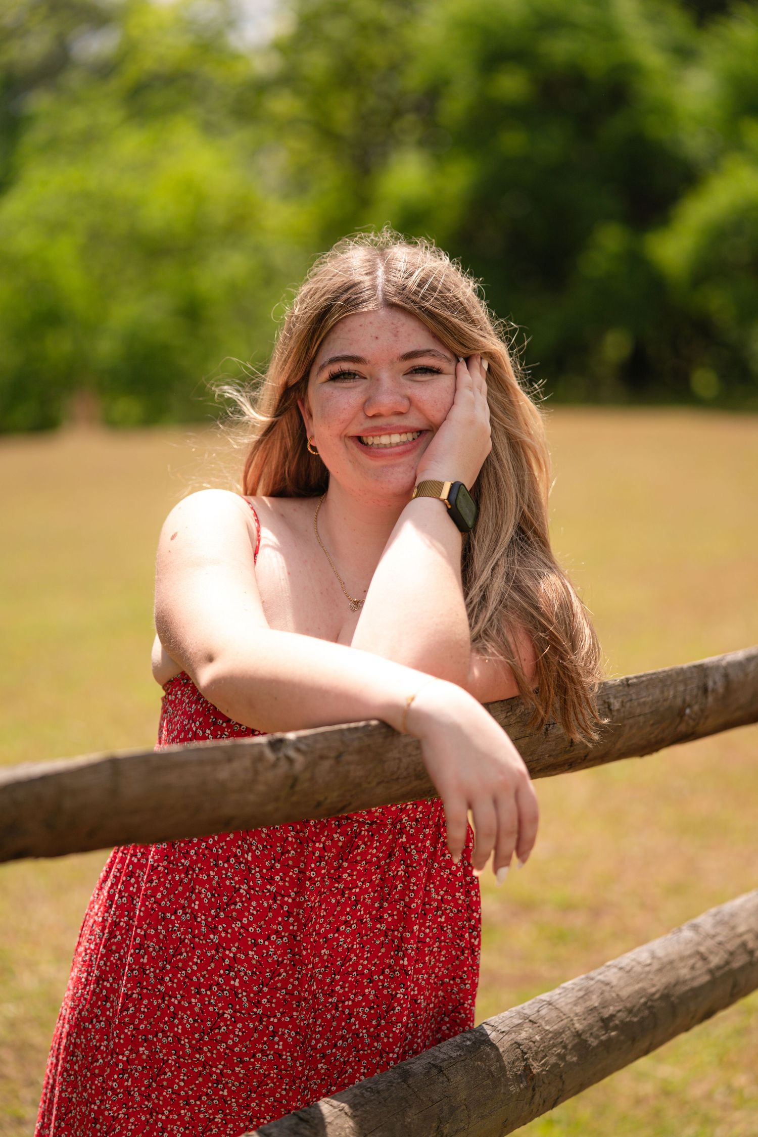 Smiling young woman in red floral dress leaning on wooden fence in sunny park setting.