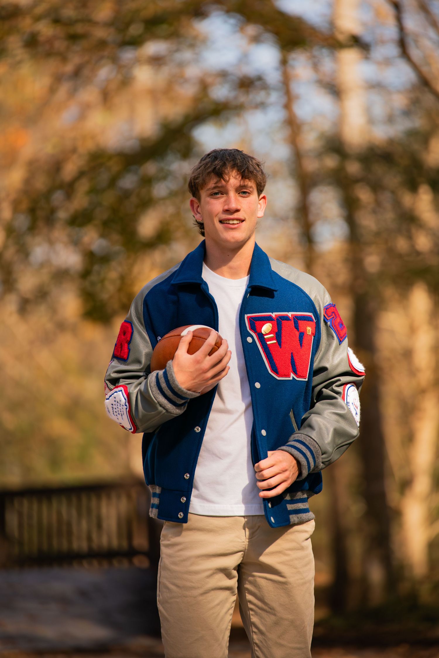 Student wearing varsity jacket holds football while posing outdoors in autumn setting.